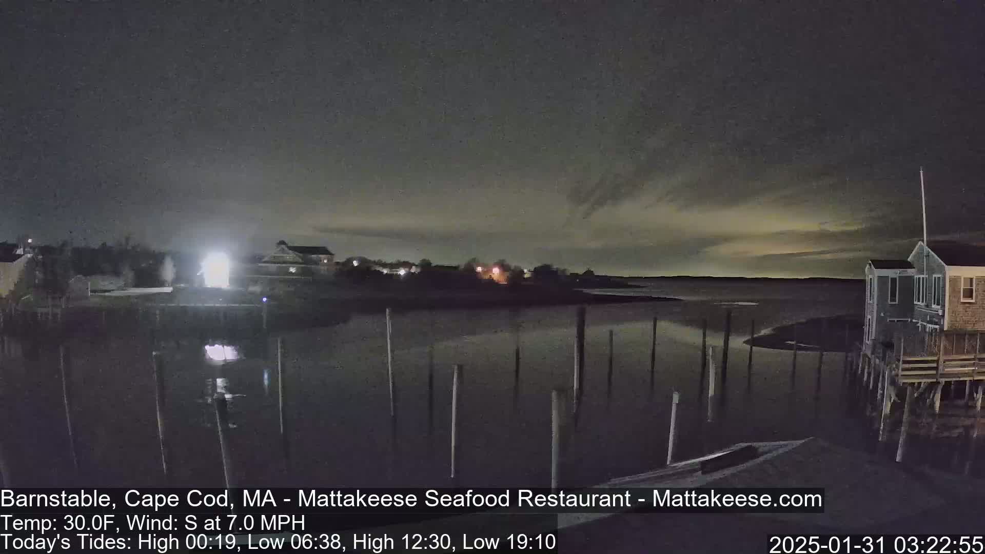 A nighttime view of a calm body of water with pilings, houses on the shore under a dark sky with faint light in the distance.