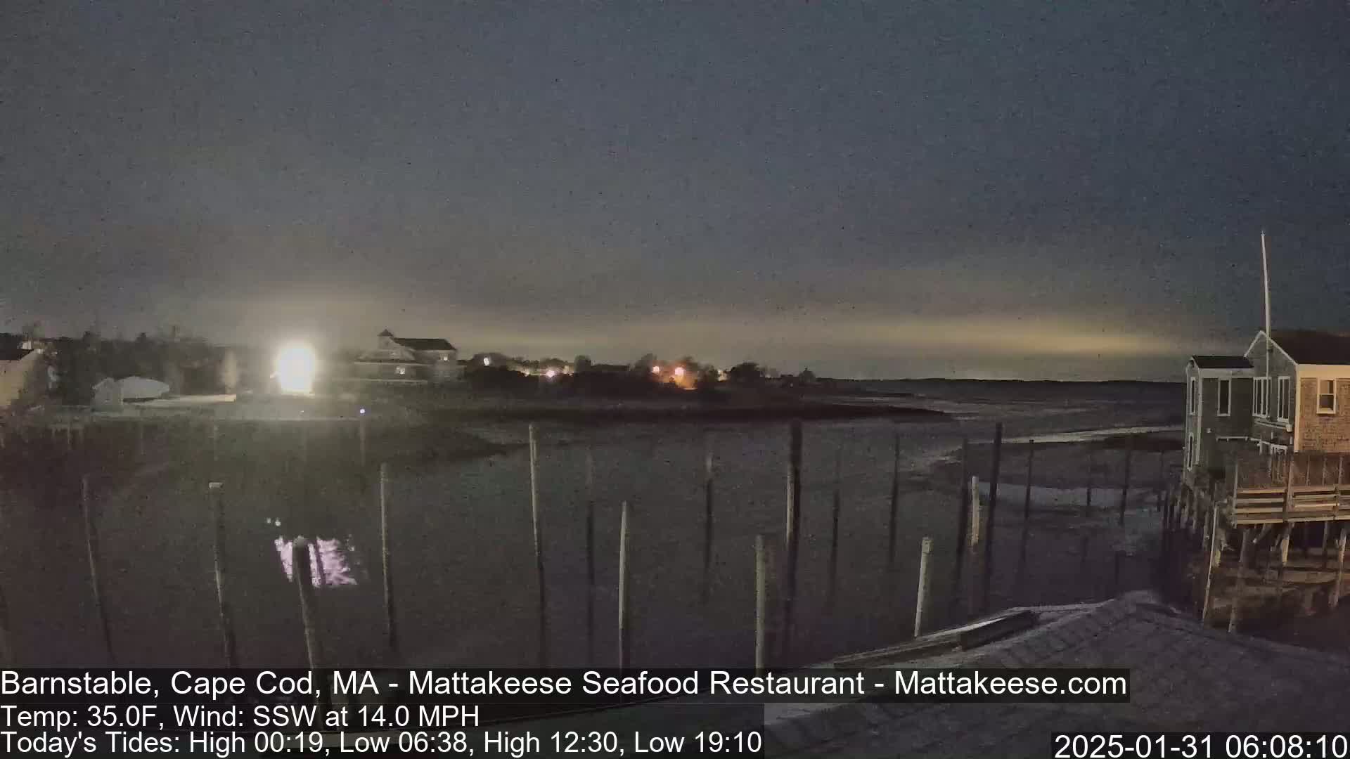 A nighttime seaside scene shows a dark, calm body of water, a few houses with lights on, and a mostly clear sky with a faint, light-colored horizon.