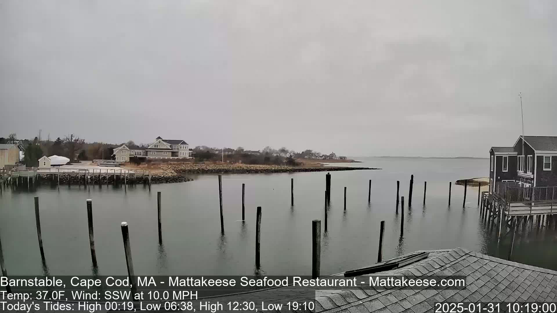 A calm, overcast day reveals a waterfront scene with a small town, a dock with pilings in the water, and a dark-colored house on pilings.