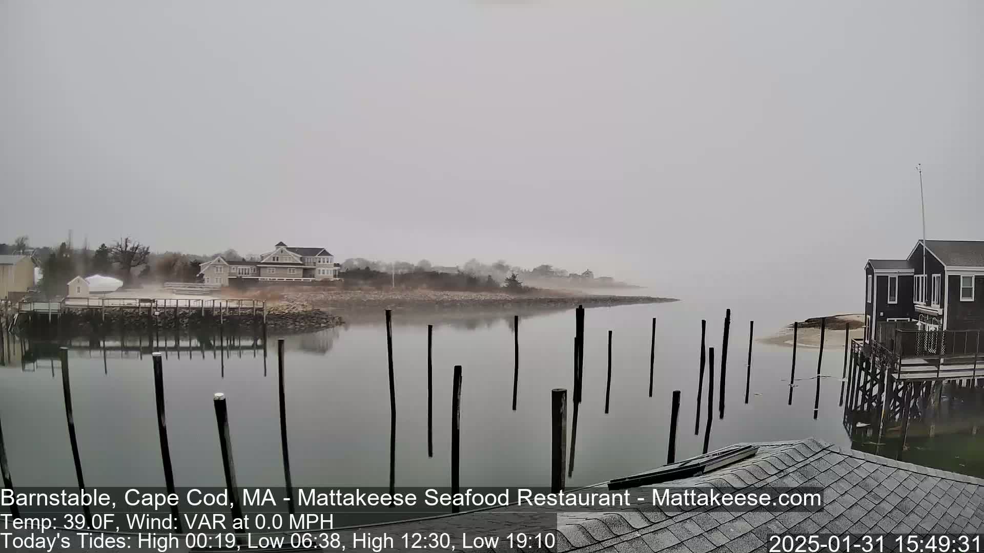 A foggy waterfront scene shows calm water with wooden posts extending from the shore, houses, and a small building near the water's edge.