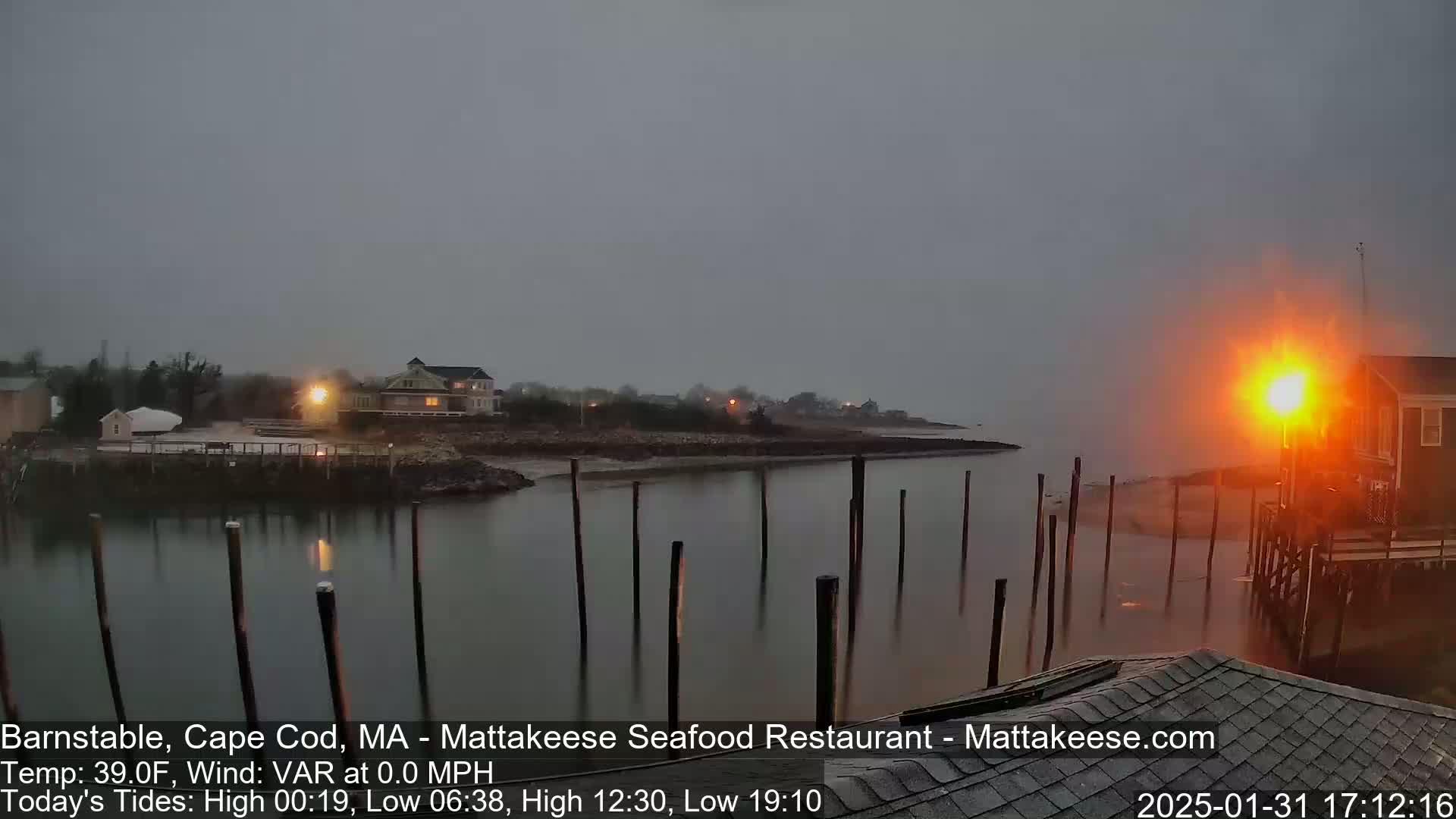 A calm, overcast harbor scene at dusk shows a waterfront with buildings, pilings in the water, and a faint orange glow from a light source.