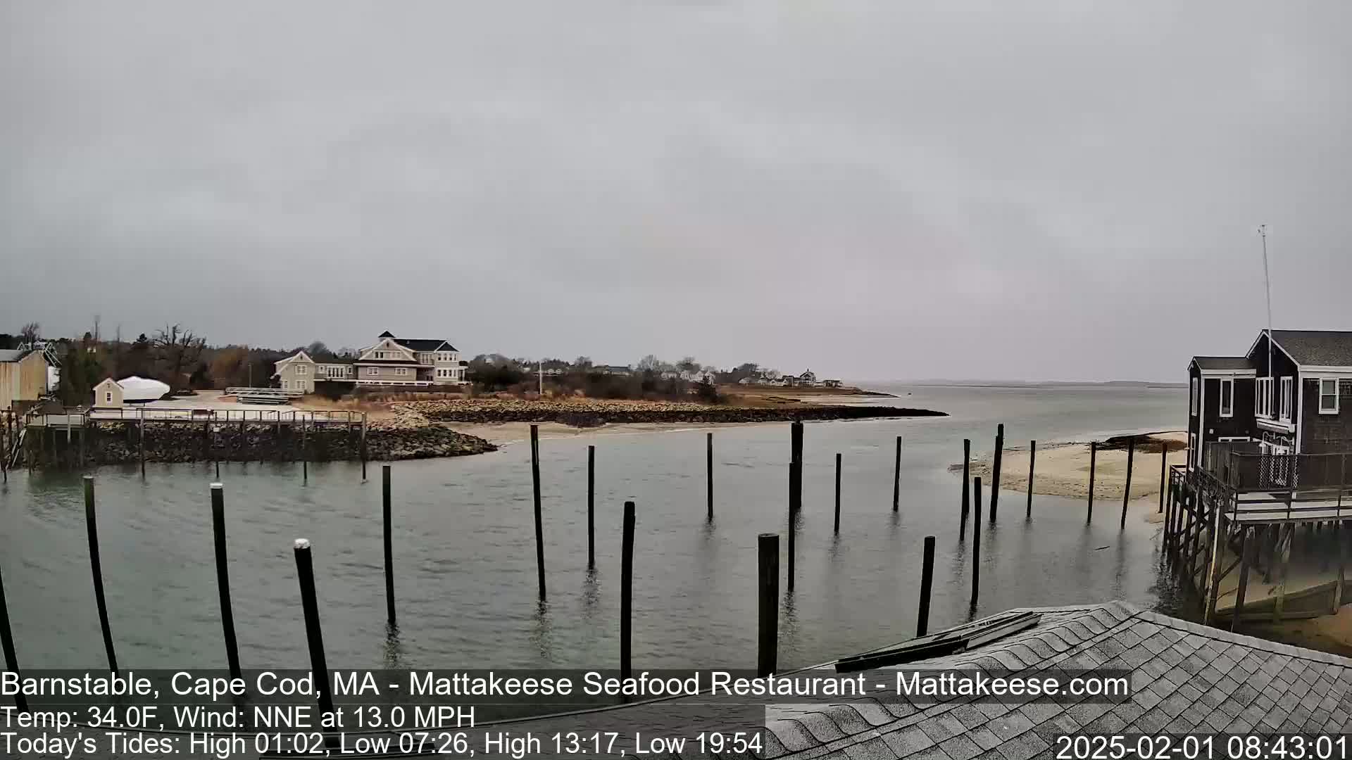 A calm body of water with wooden pilings extends from a shoreline featuring houses and a gray overcast sky.