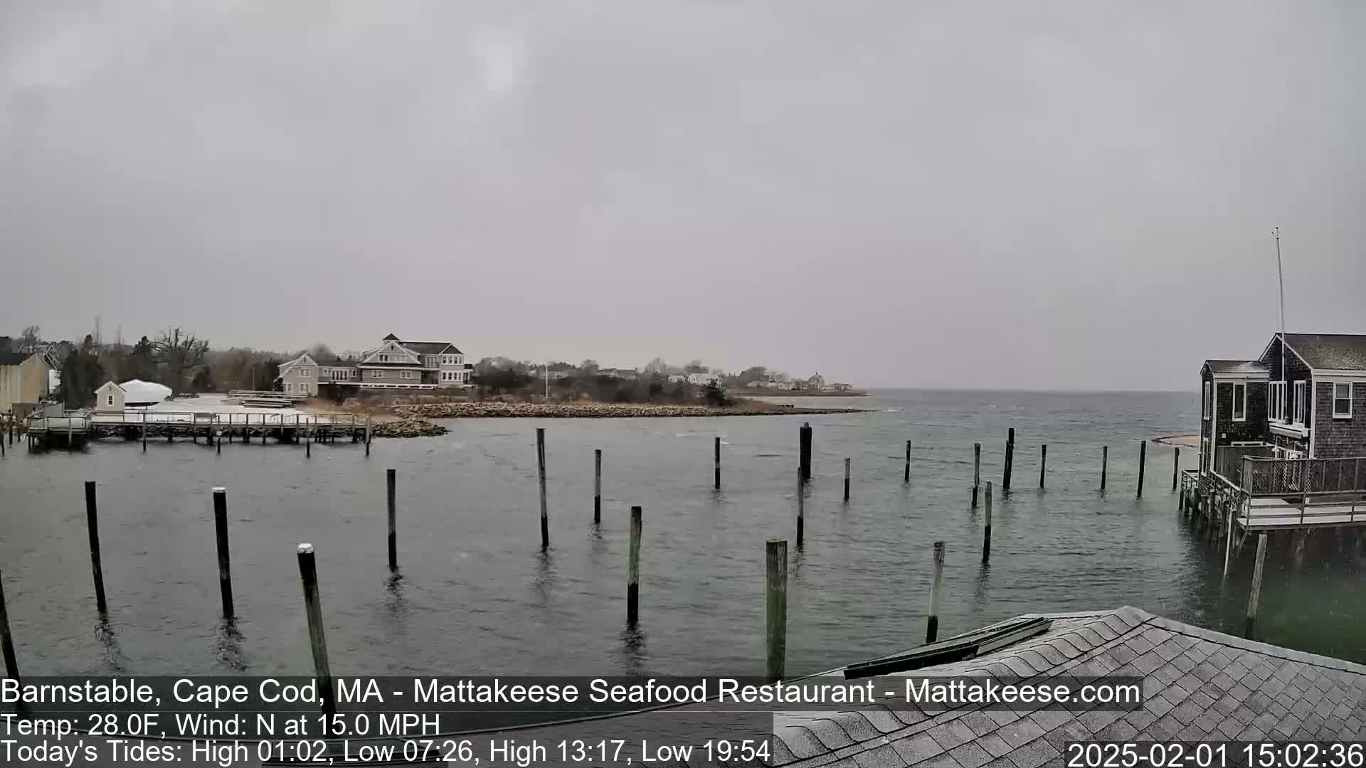 A snow-covered waterfront scene shows a calm body of water with several wooden pilings, a dock with buildings, and houses along the shore under an overcast sky.