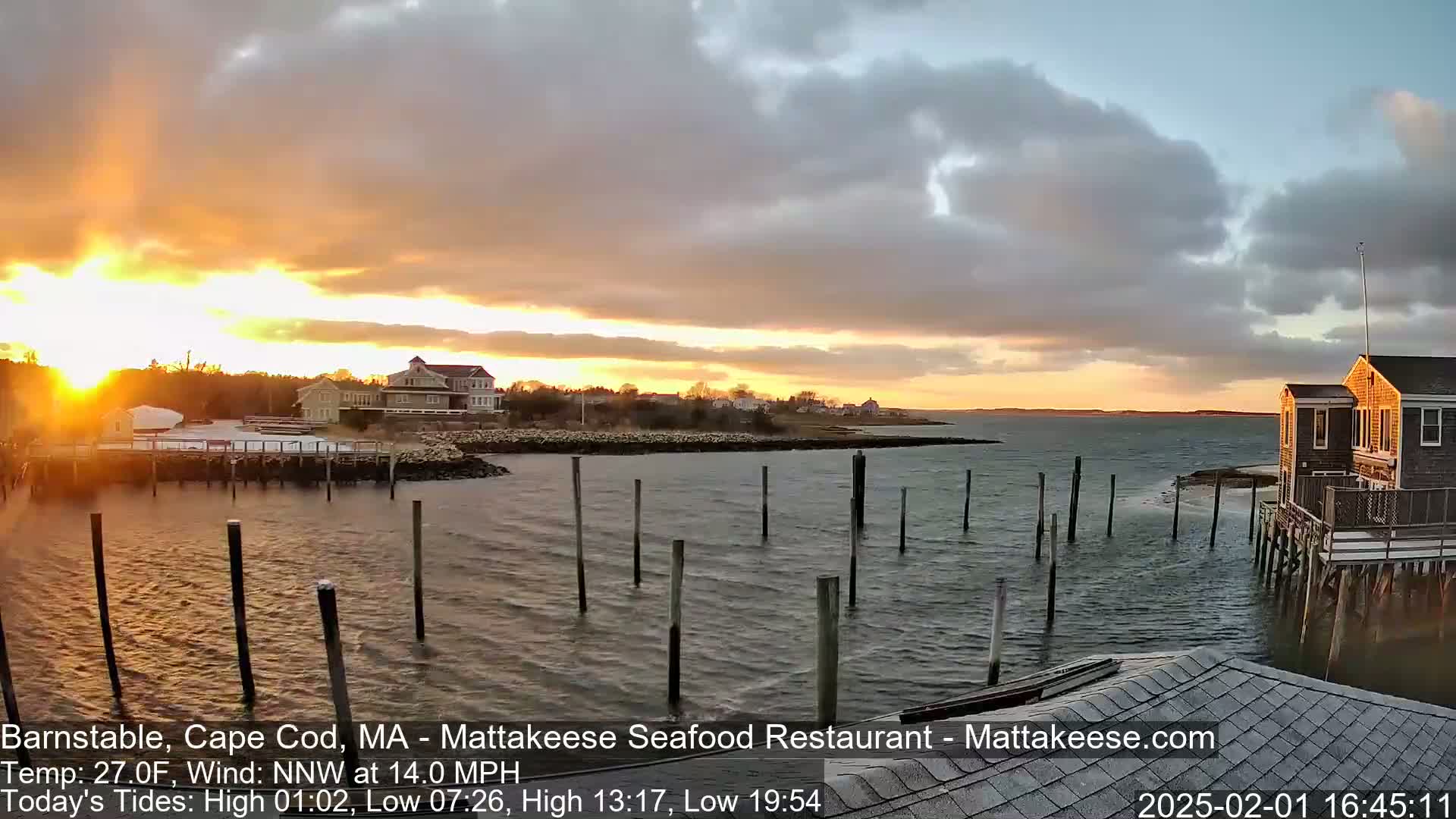 A sunset over a calm bay with a waterfront village and numerous pilings in the water, under a partly cloudy sky.