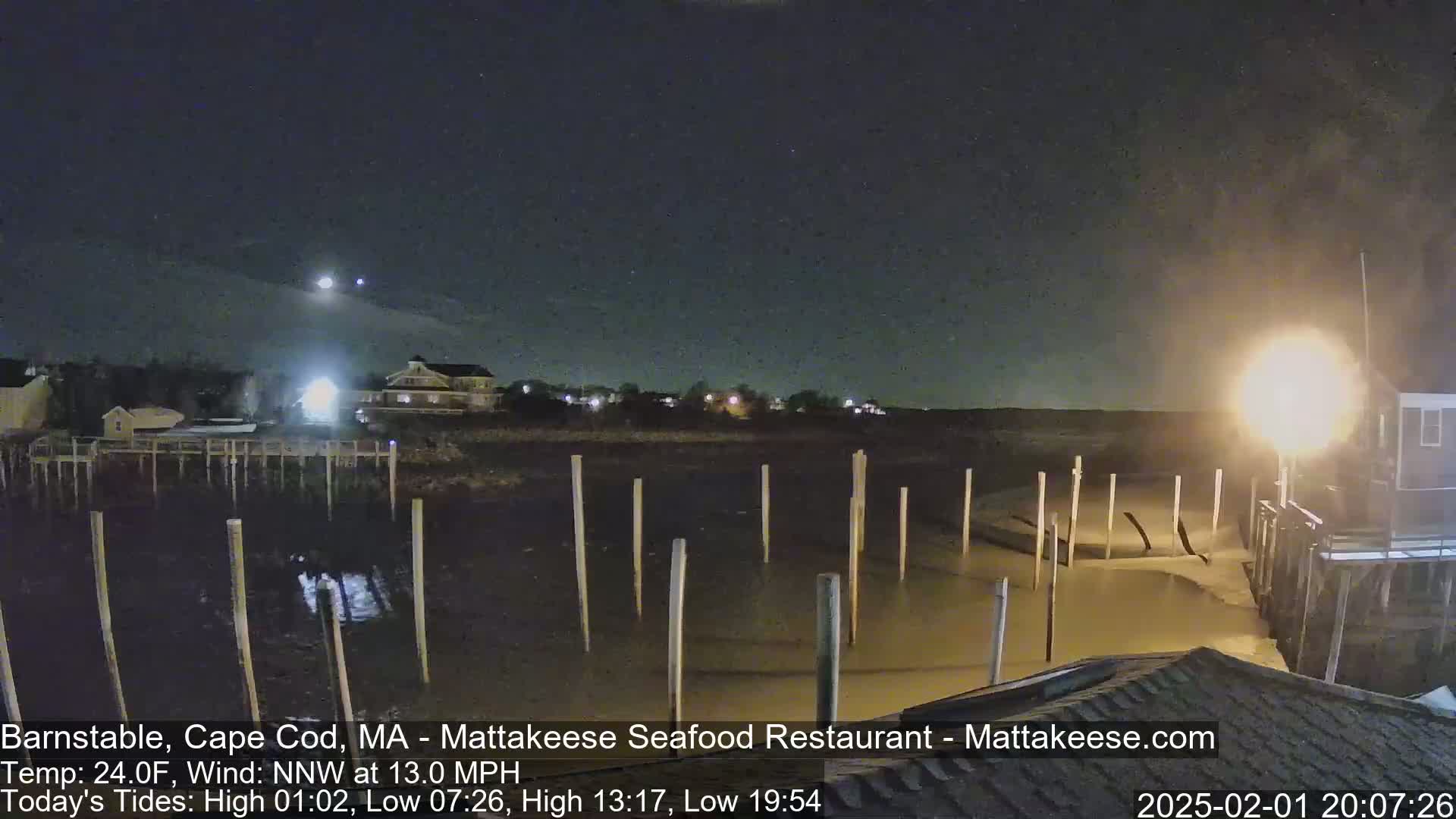 A nighttime view of a calm body of water with wooden pilings, illuminated by lights from nearby buildings under a clear, starry sky.