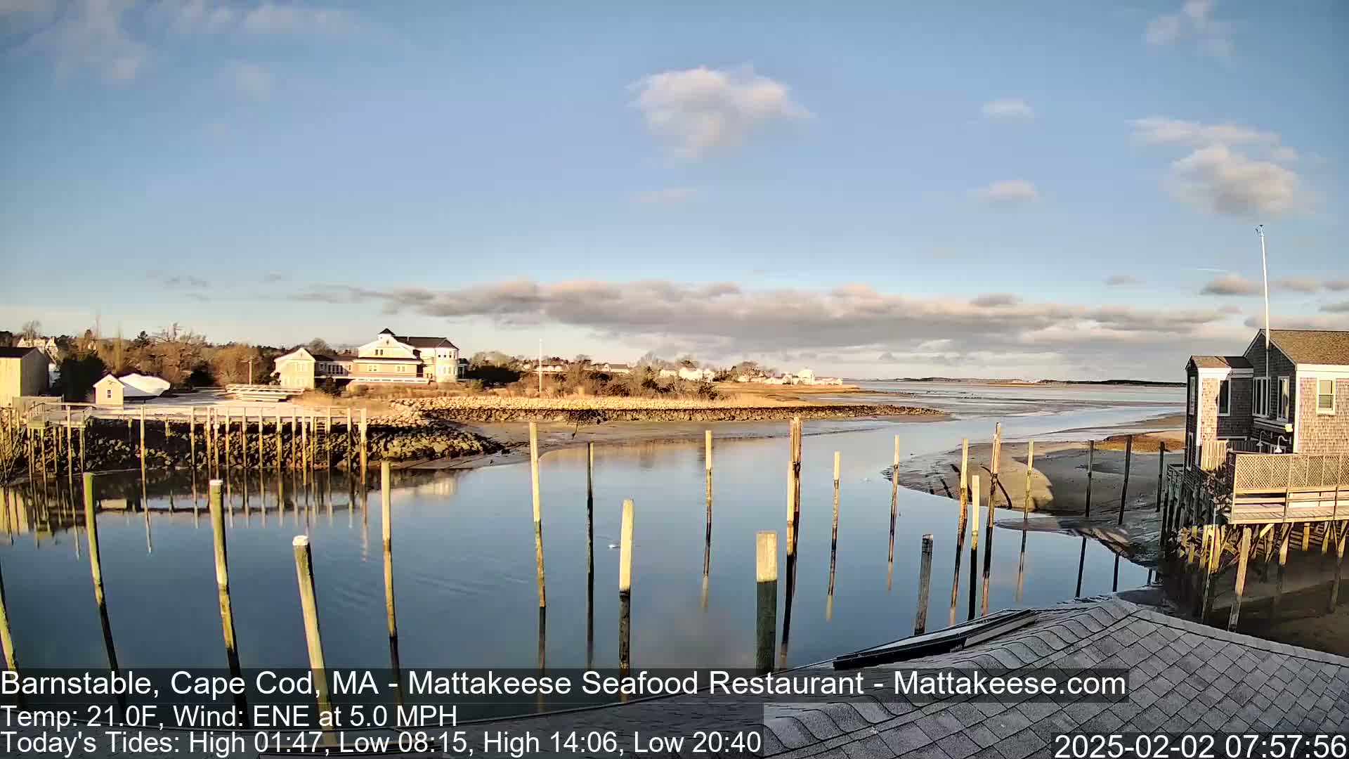 A calm, partly cloudy day reveals a waterfront scene with buildings, a calm waterway, and wooden pilings extending into the water.