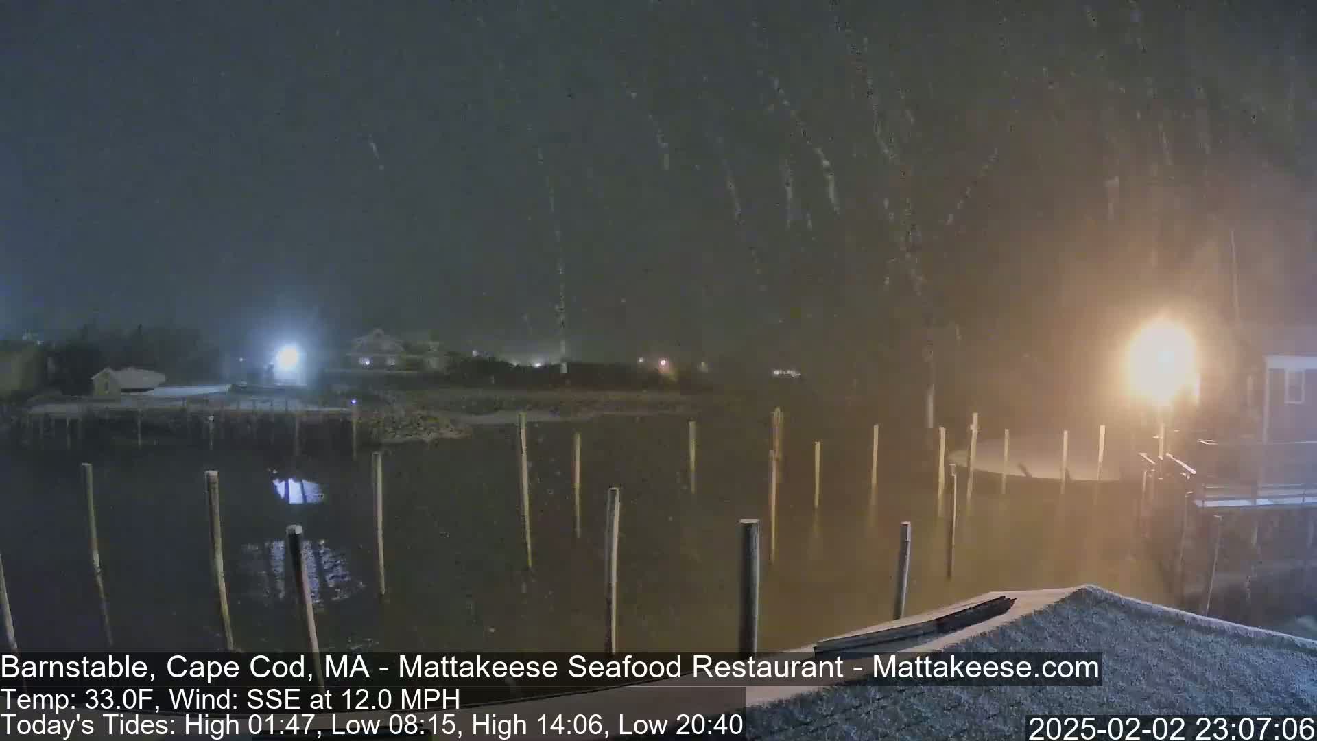 A nighttime scene of a snow-covered waterfront with several pilings extending into calm water, illuminated by nearby lights.