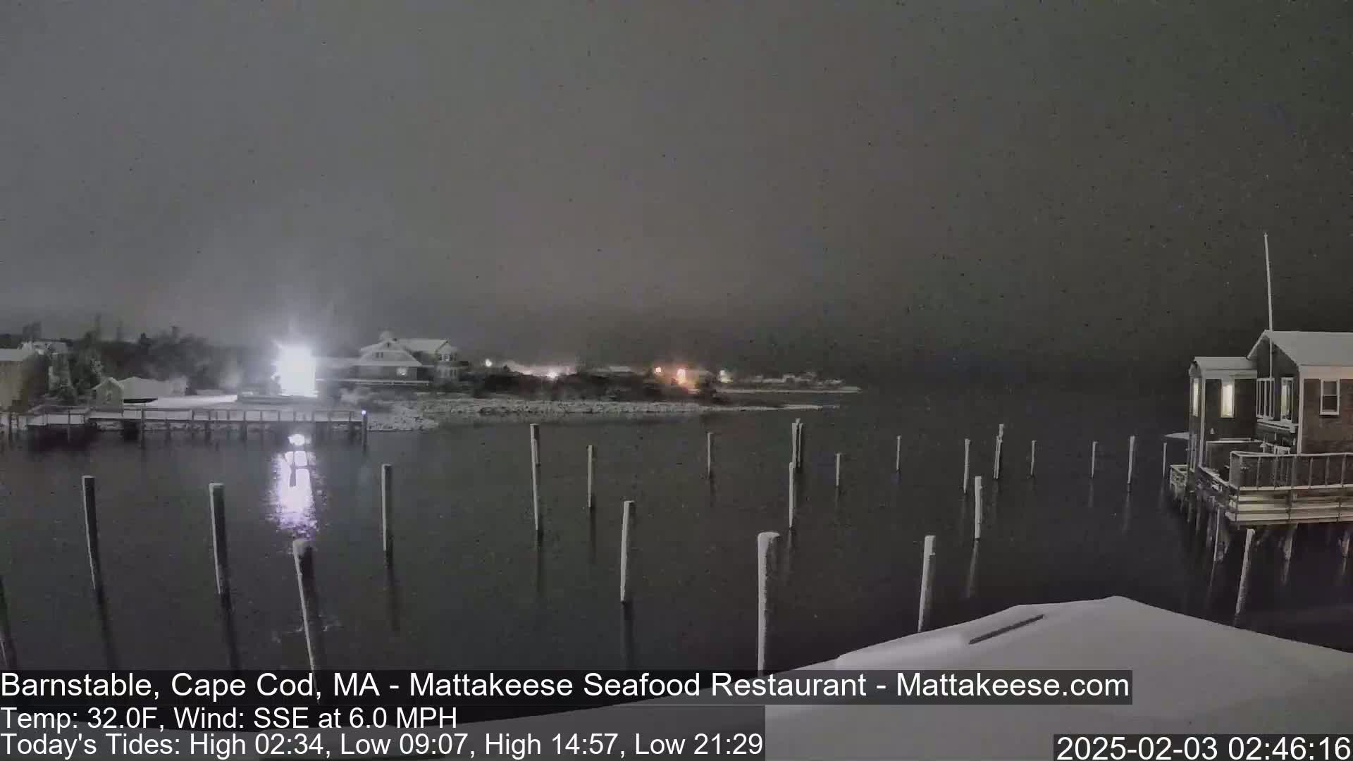 A nighttime snow scene shows a body of water with wooden pilings, snow-covered buildings on the shore, and lightly falling snow.
