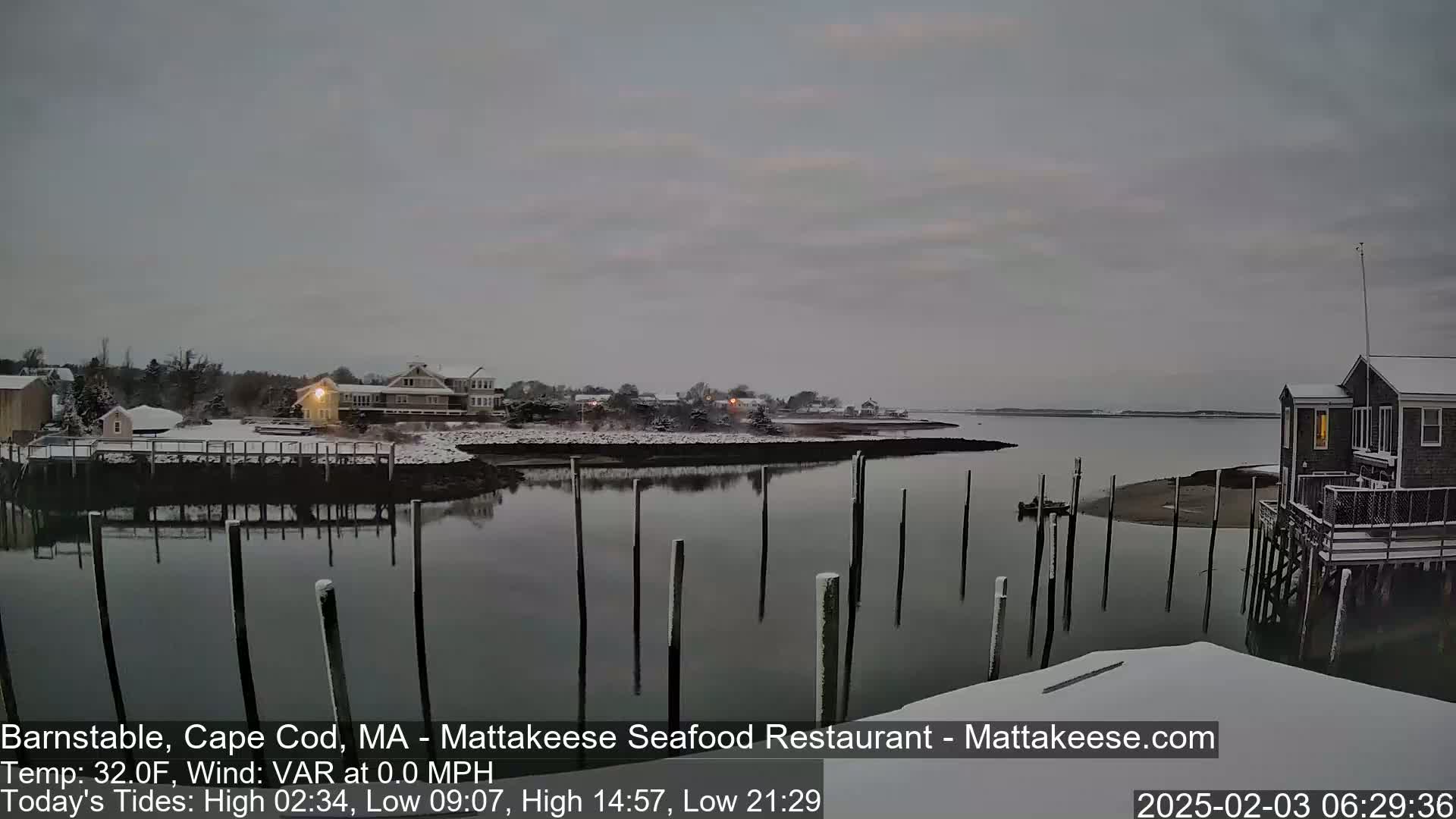 A snow-covered waterfront scene shows calm water reflecting a gray sky, with several buildings and wooden pilings in the foreground.