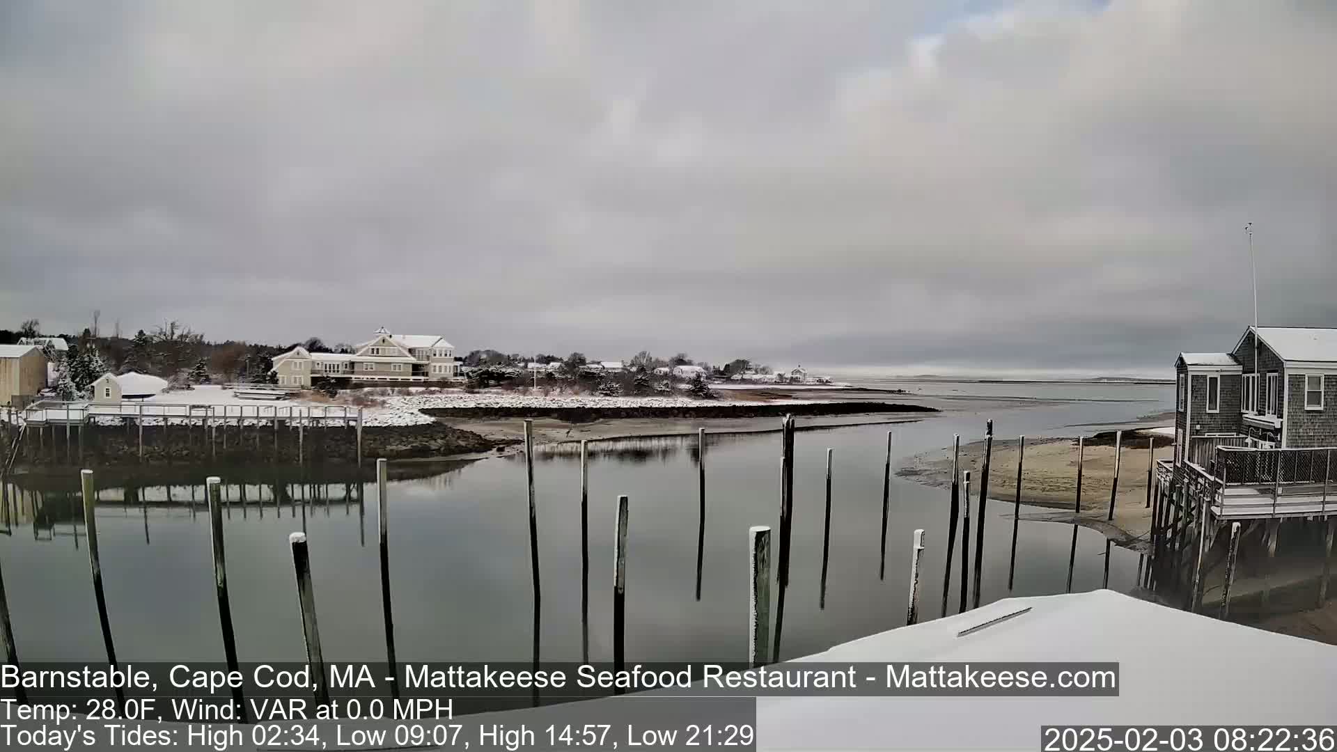 A snow-covered waterfront scene shows calm water reflecting a cloudy sky, with several buildings and weathered wooden pilings in the foreground.