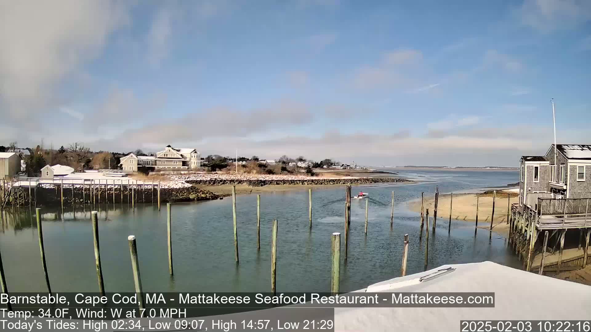 A calm waterway with wooden pilings and a small boat, nestled between a snow-dusted shoreline and buildings under a partly cloudy sky.