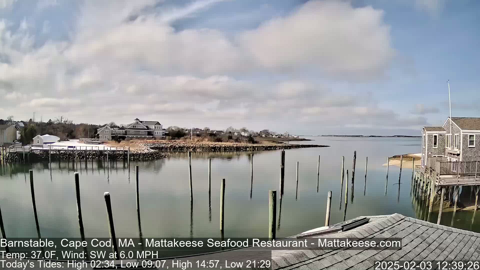 A calm body of water with wooden pilings extends from a shoreline of houses and a sandy beach under a partly cloudy sky.