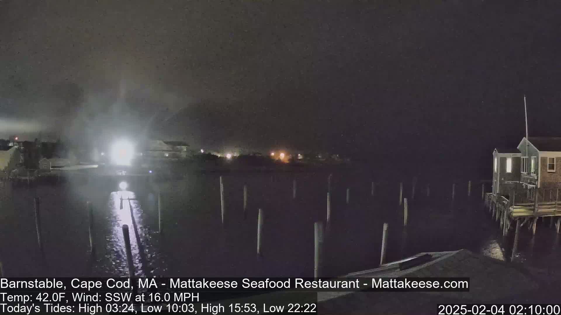 A nighttime view of a dark body of water with pilings, a building on stilts, and lights in the distance, under a cloudy sky with a 16 mph southwest wind.