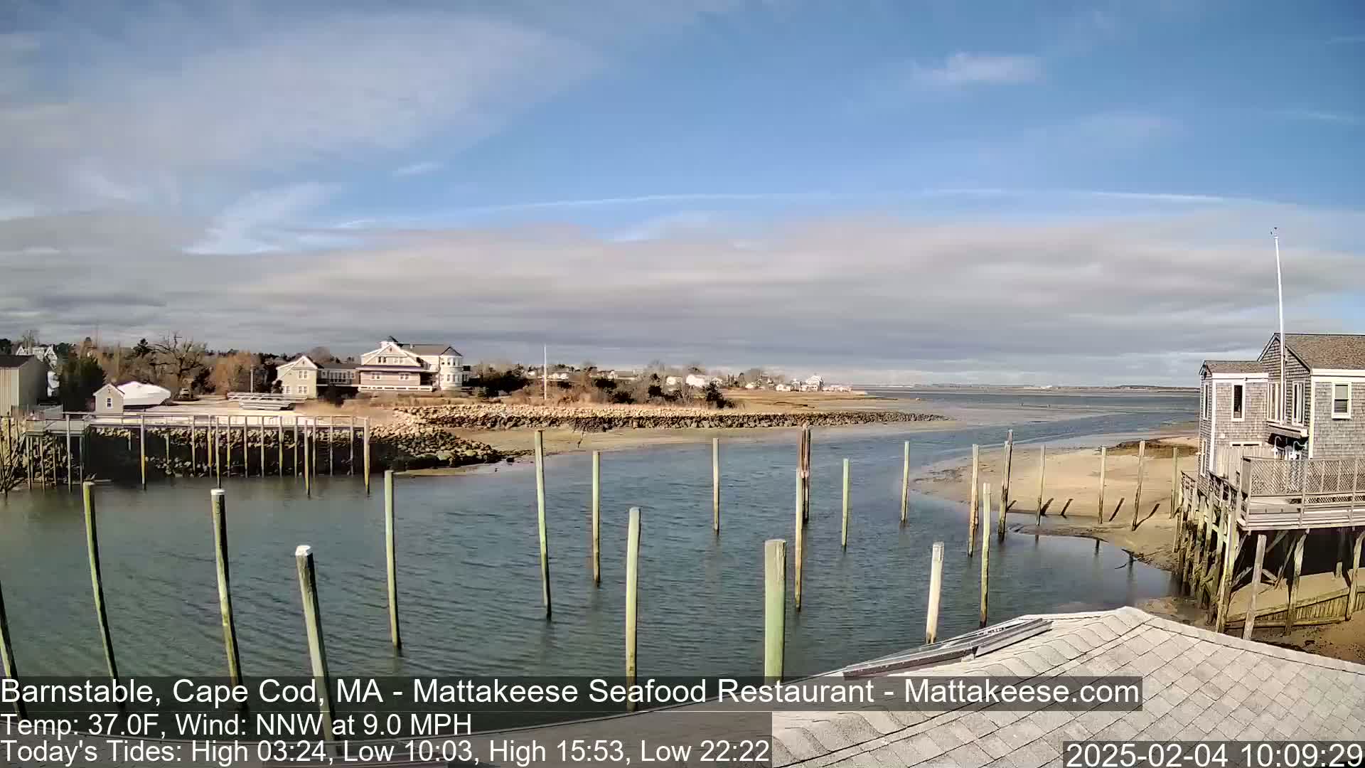 A calm, partly cloudy day shows a waterfront scene with a narrow waterway, wooden pilings, houses, and a sandy shore.