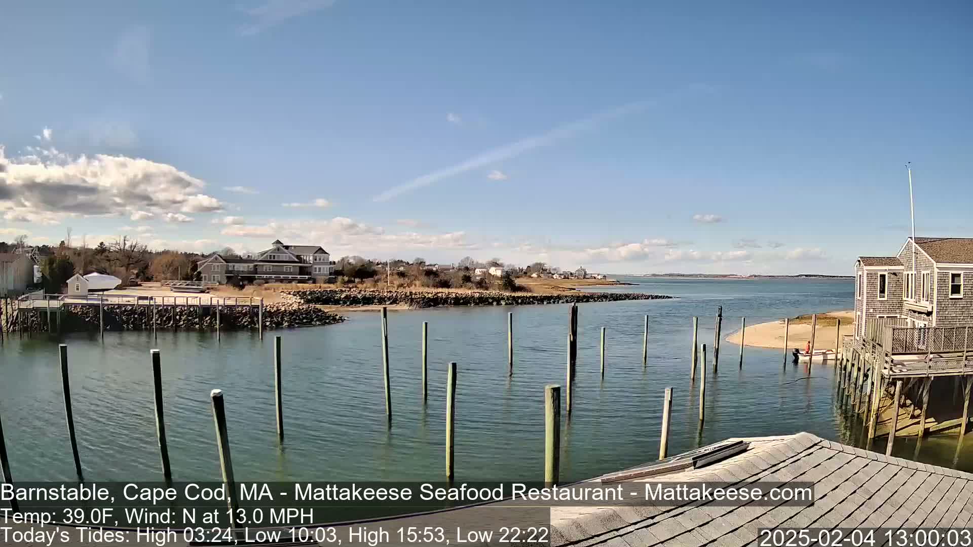 A calm waterway with pilings, houses, and a sandy shore under a partly cloudy sky.