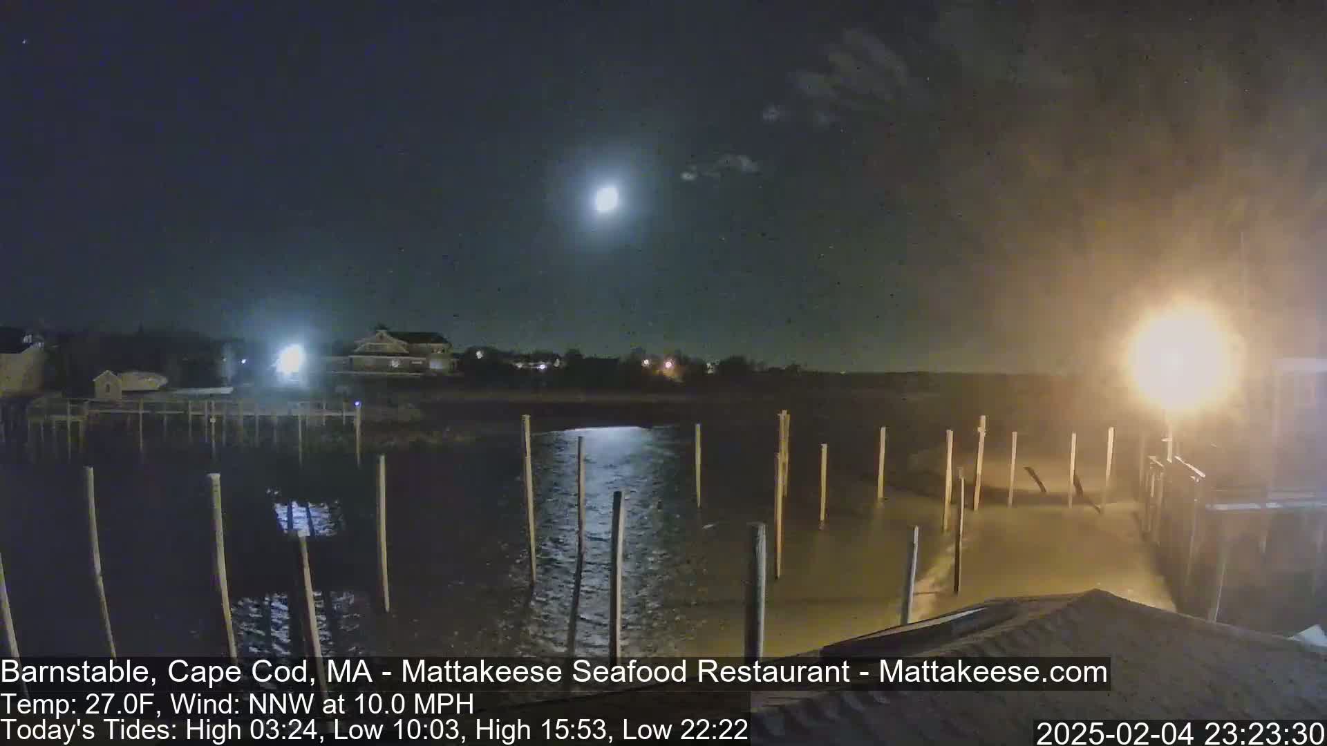 A nighttime view of a calm body of water with wooden pilings, a building in the background under a bright moon and partly cloudy sky.