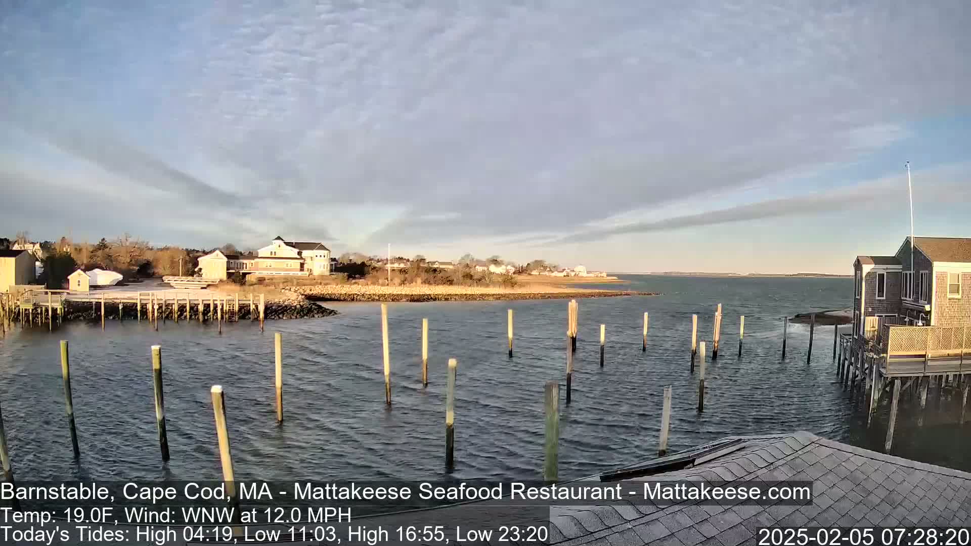 A partly cloudy, cool day shows a waterfront scene with a calm body of water, wooden pilings in the water, buildings on the shore, and a few small boats.