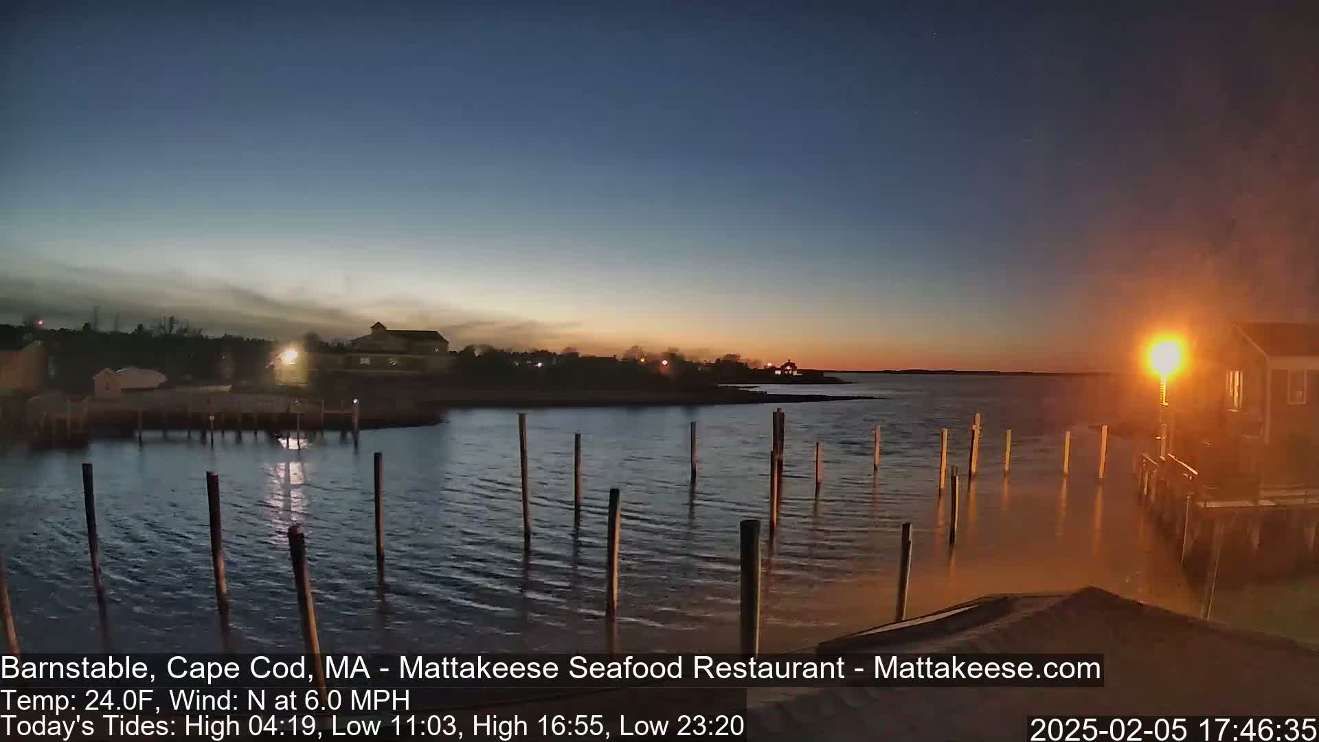 A calm body of water at sunset, with a few lights on the shore and several posts sticking up from the water.