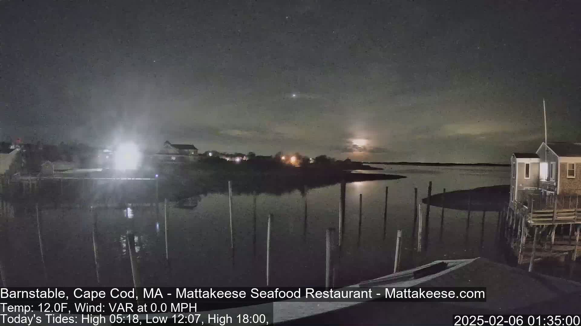 A nighttime view of a calm body of water with a few lights on the shore under a partly cloudy sky, with a bright moon visible.