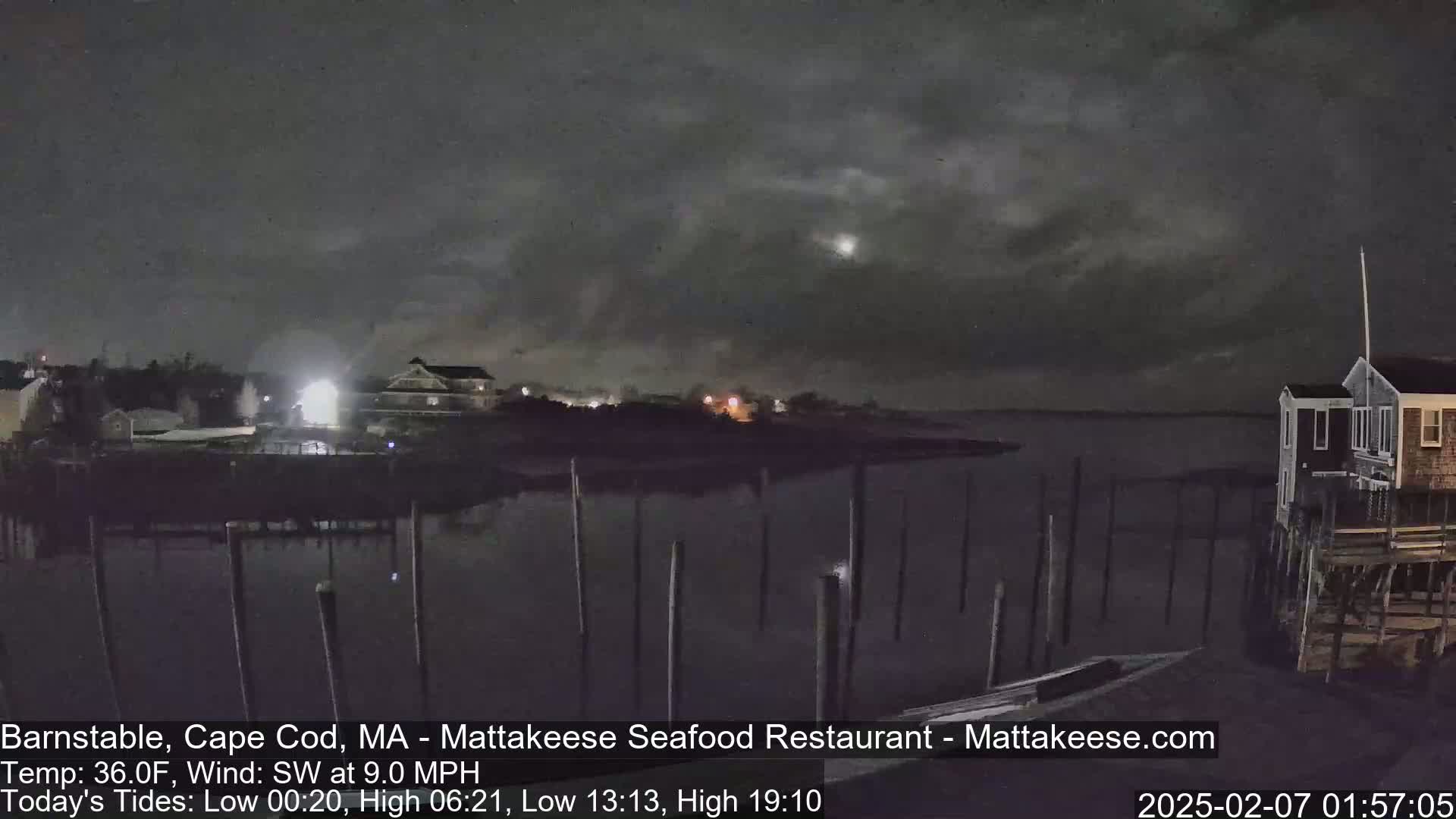 A nighttime view of a calm body of water with a few buildings and pilings visible under a dark, cloudy sky.