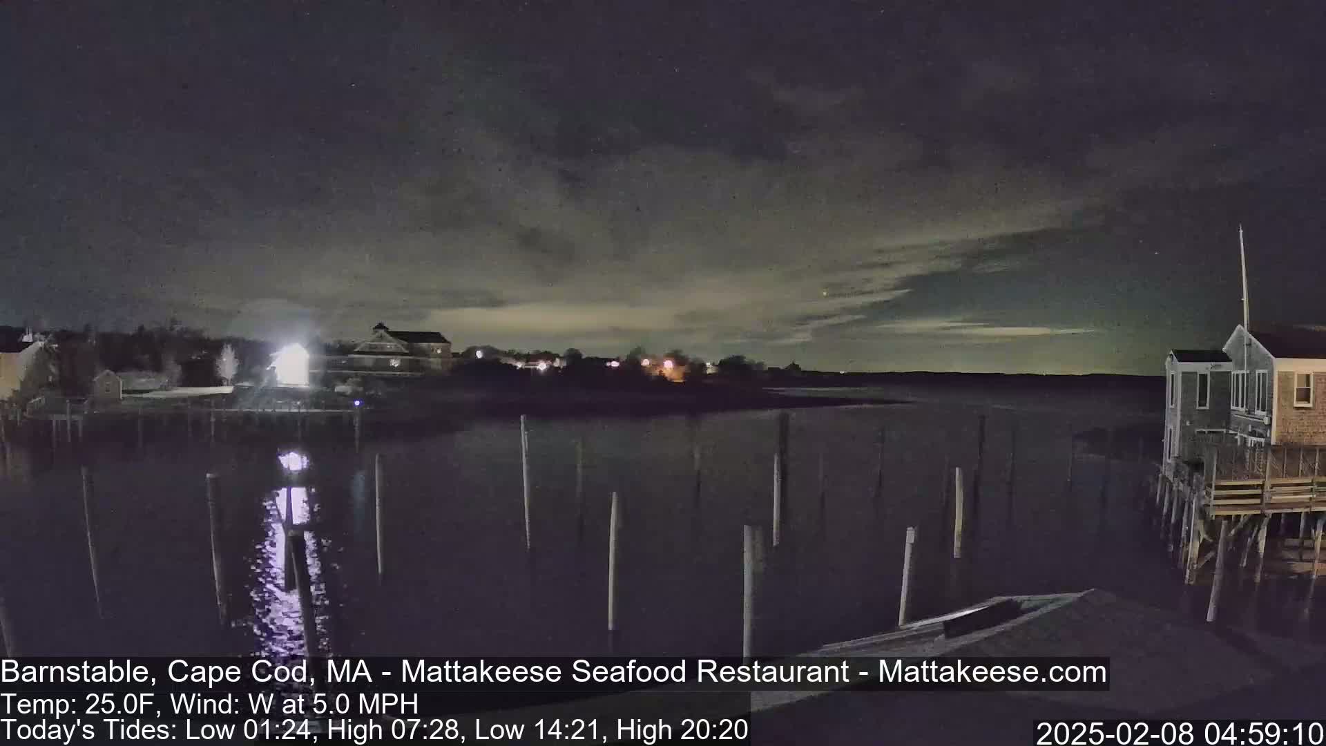 A nighttime view of a calm body of water with pilings, buildings, and trees along the shore under a mostly clear, dark sky.