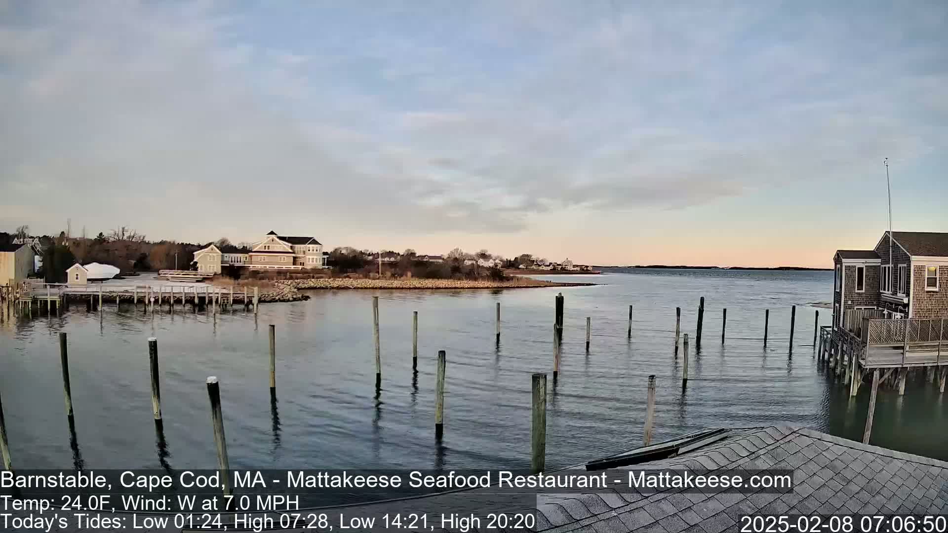 A calm body of water with wooden pilings extends from a shoreline with houses and low-lying vegetation under a partly cloudy sky.