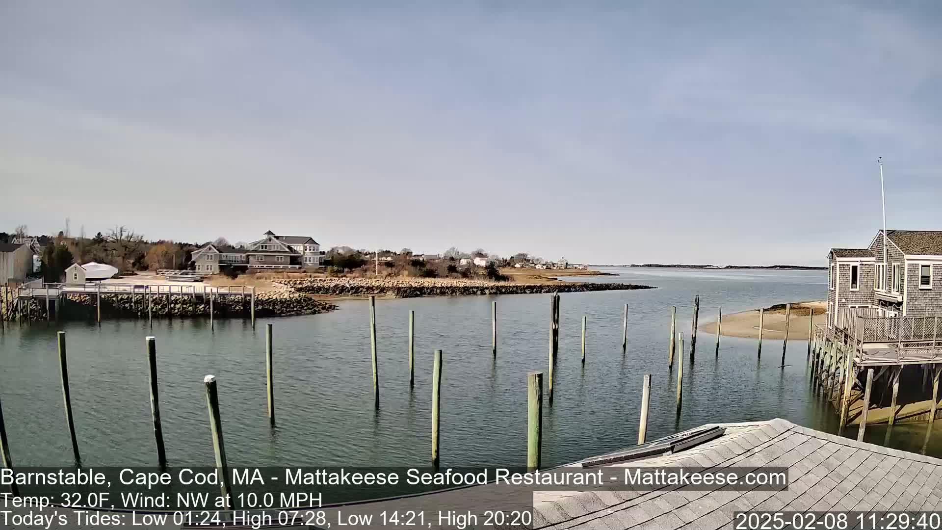A calm body of water with wooden pilings extends from a shoreline featuring houses and a small beach, under a clear, bright sky.