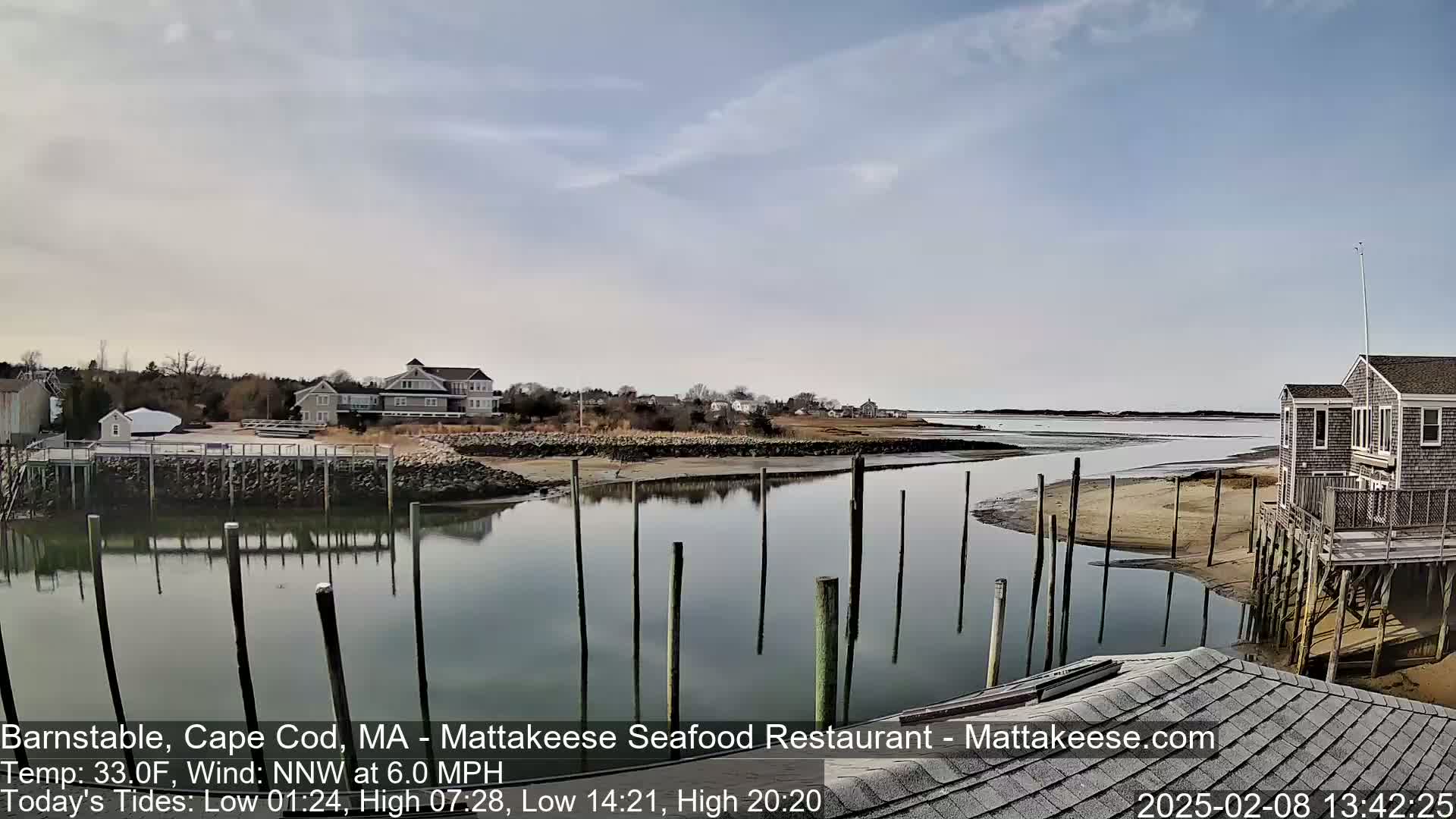 A calm waterway reflects houses and a partly cloudy sky, with a few weathered pilings in the water.