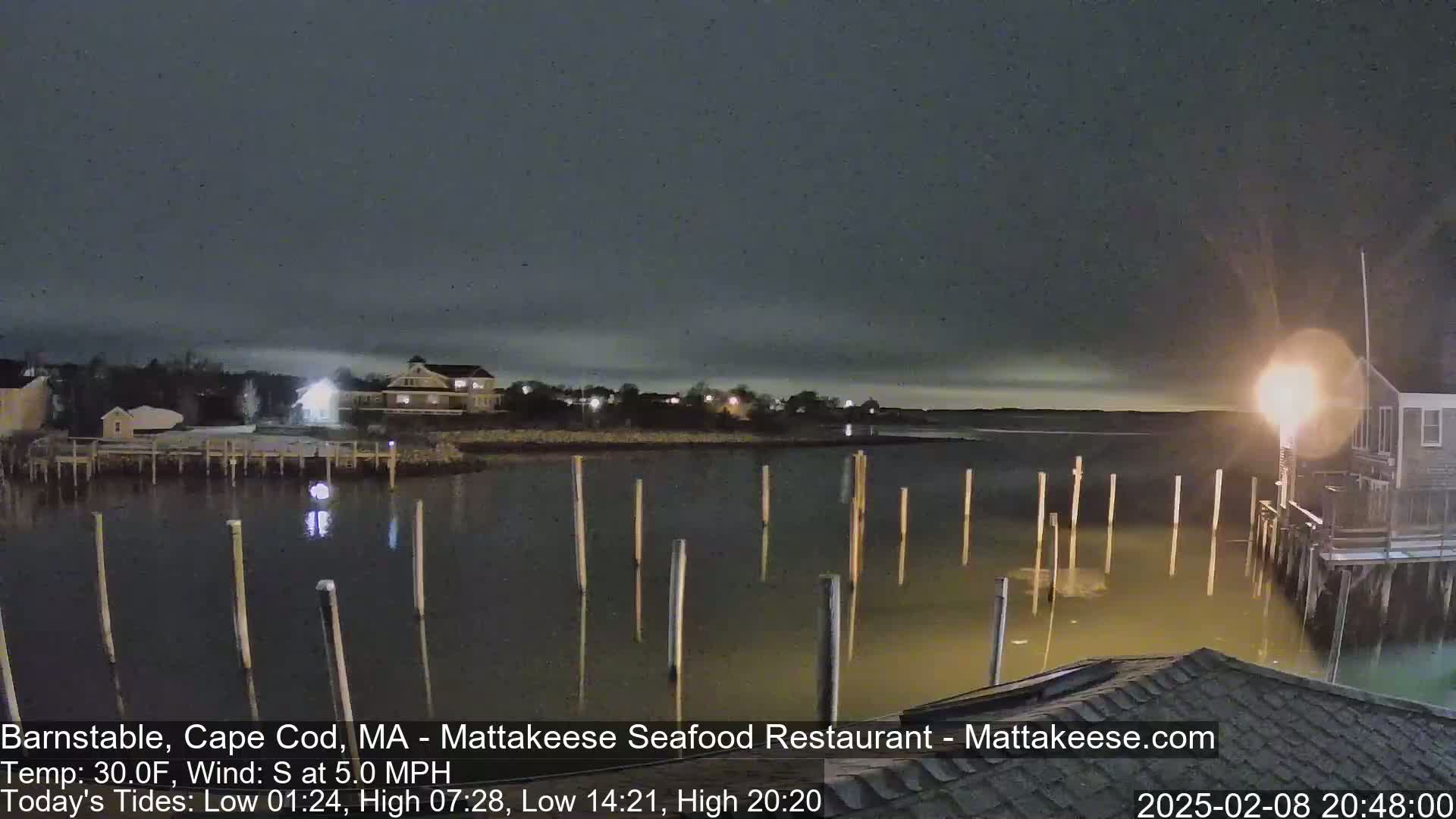 A nighttime view of a calm body of water with several pilings and waterfront buildings under a dark, cloudy sky.