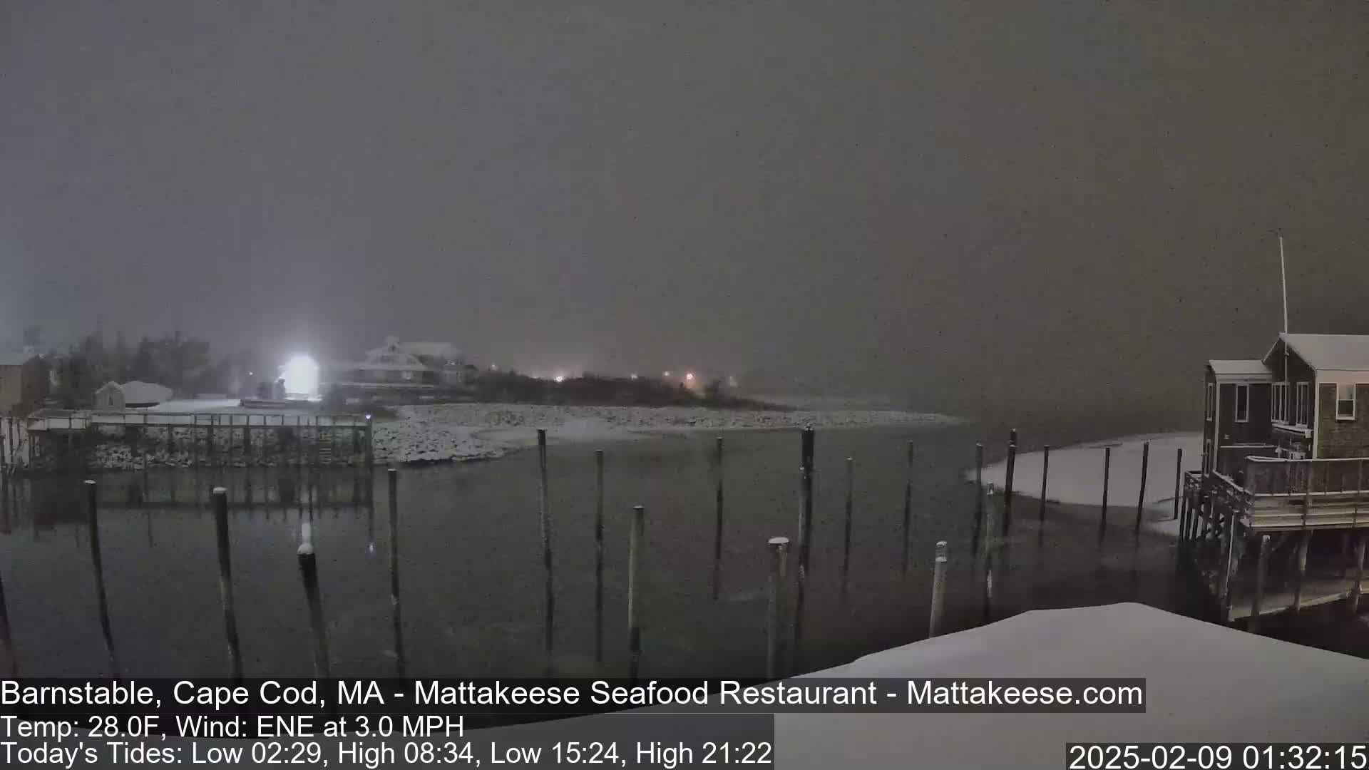 A snow-covered waterfront scene at night shows a dark body of water with pilings, and buildings under a lightly snowing sky.