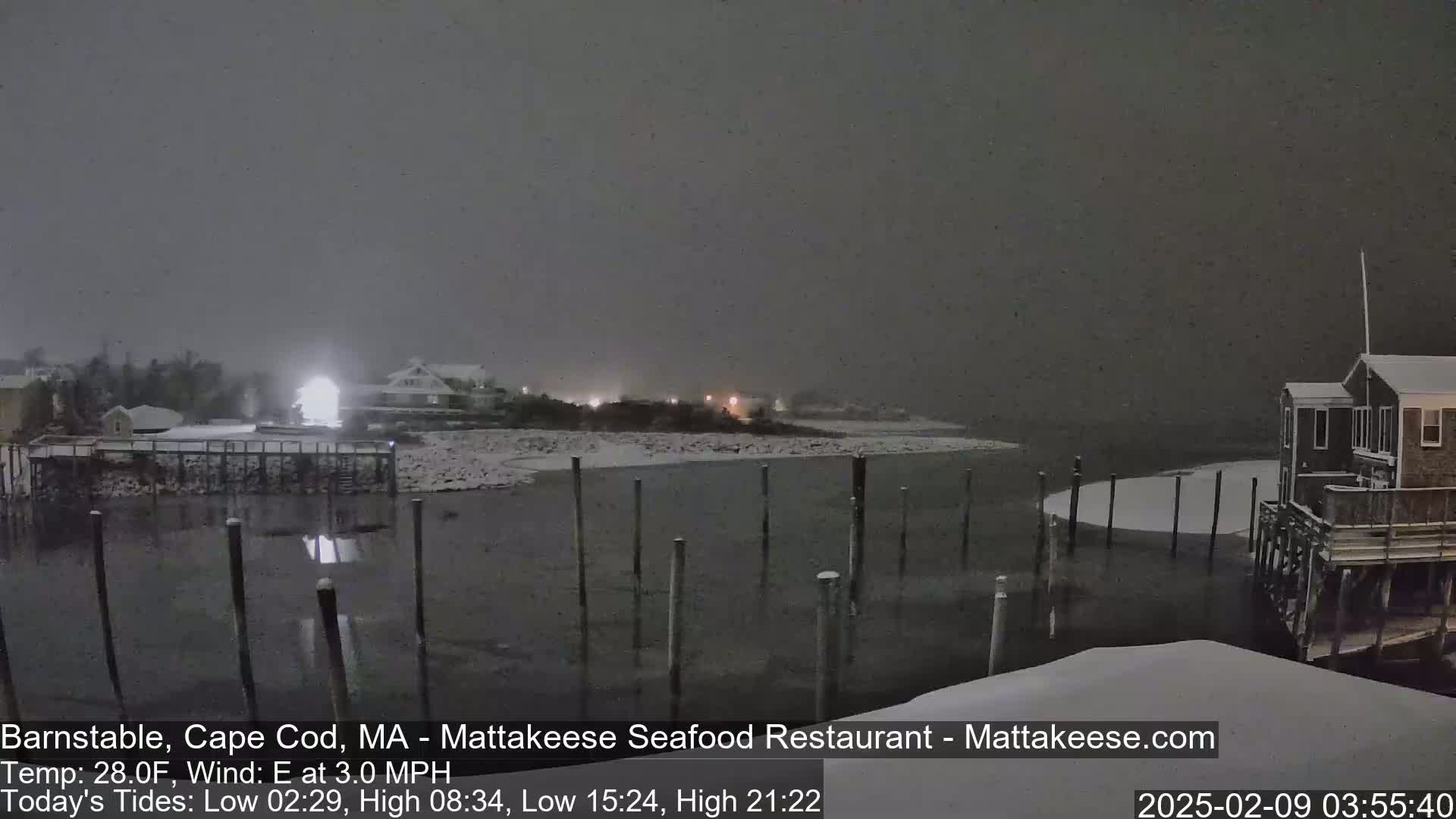 A snow-covered waterfront scene at night shows a calm body of water with pilings, several buildings, and a lightly snow-covered shoreline.