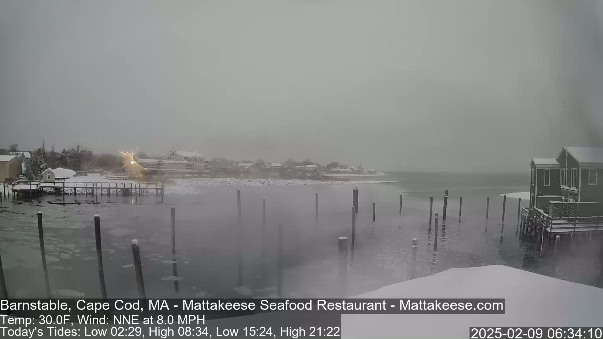 A snow-covered waterfront scene shows a calm body of water with wooden pilings, several snow-covered buildings, and a light dusting of snow in the air.