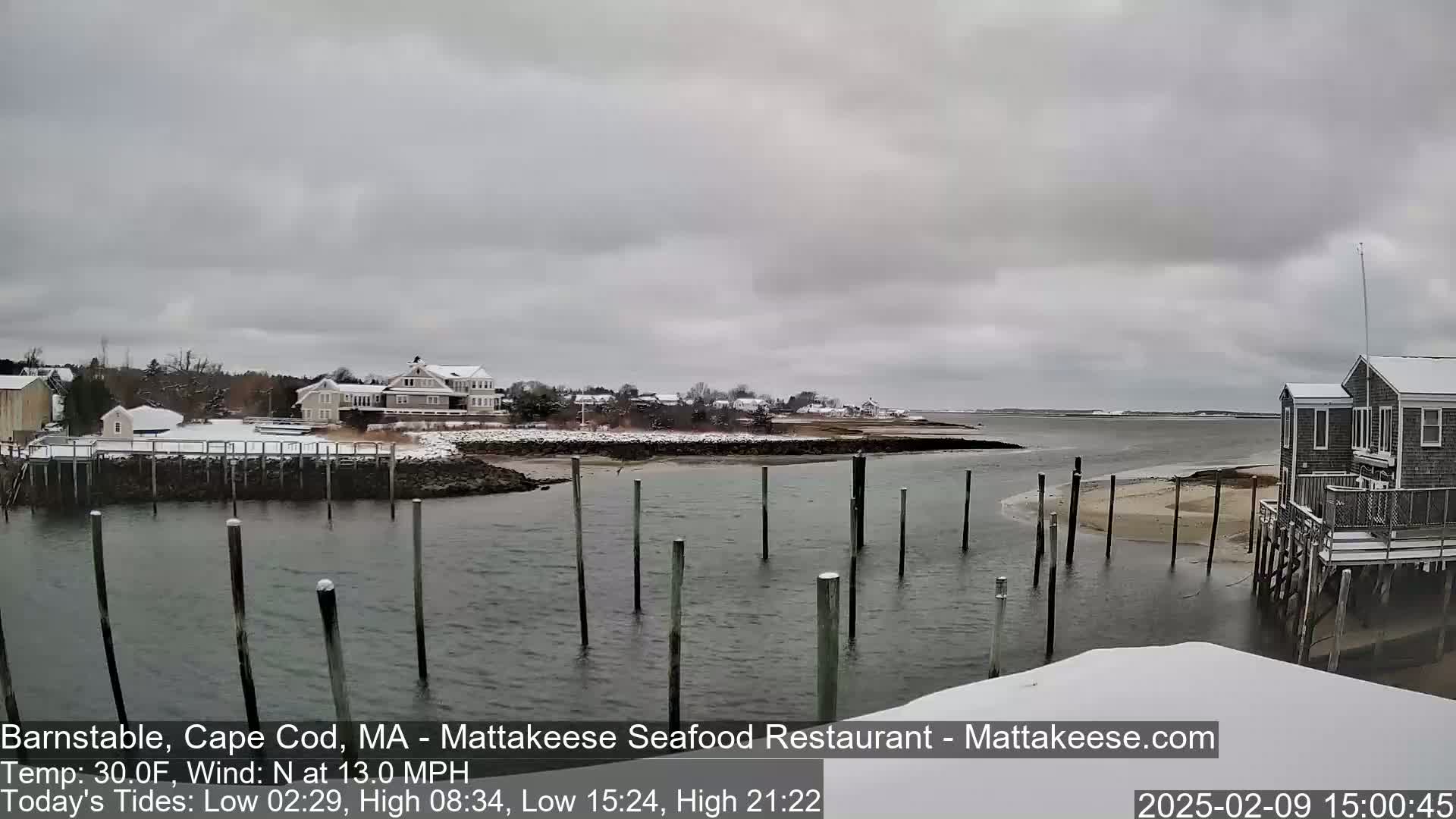 A snow-covered waterfront scene shows a calm body of water with wooden pilings, several buildings, and a cloudy sky.