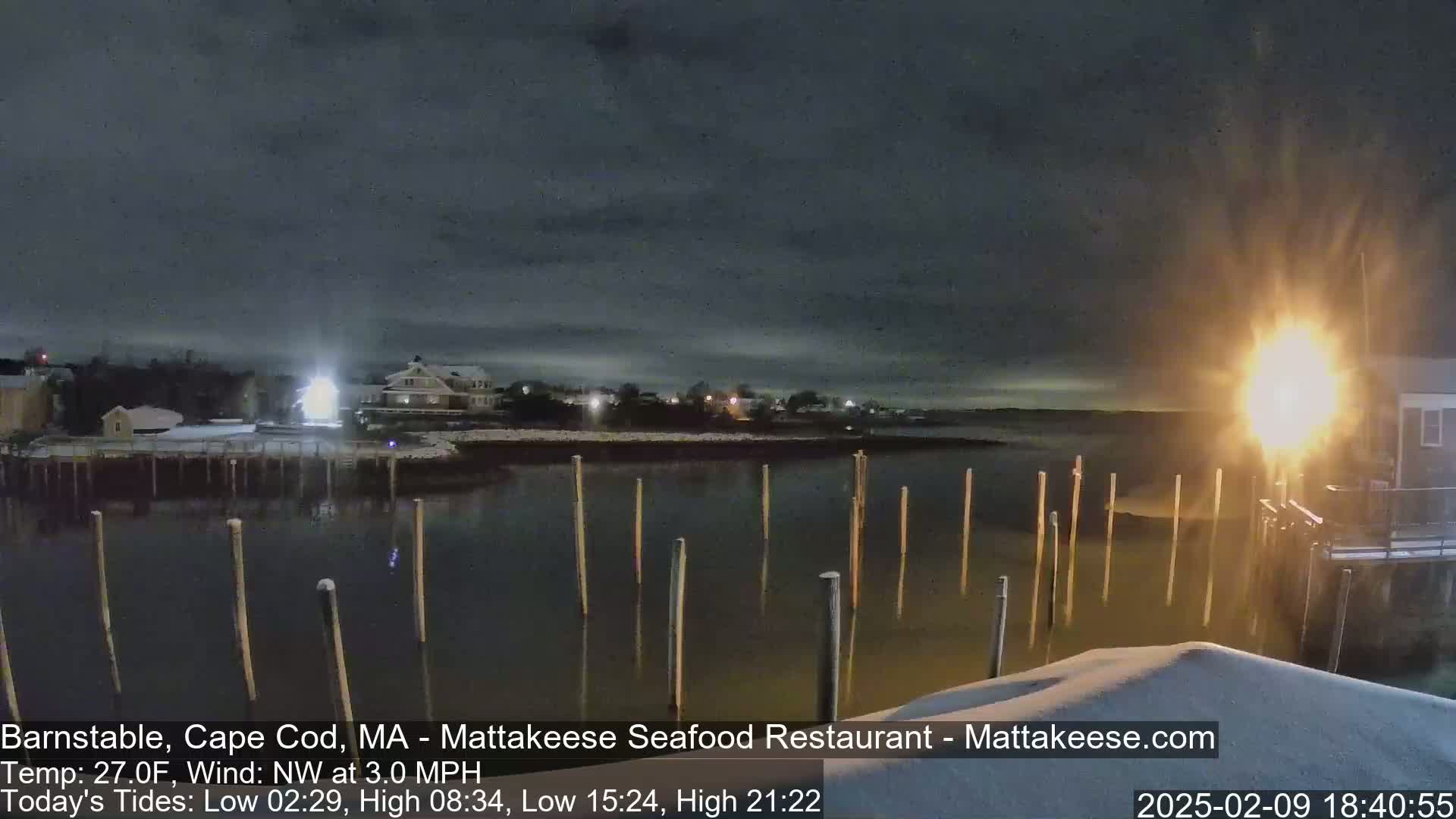 A nighttime view of a snow-covered waterfront scene with calm water, wooden posts in the water, and a few buildings in the background under a dark, cloudy sky.