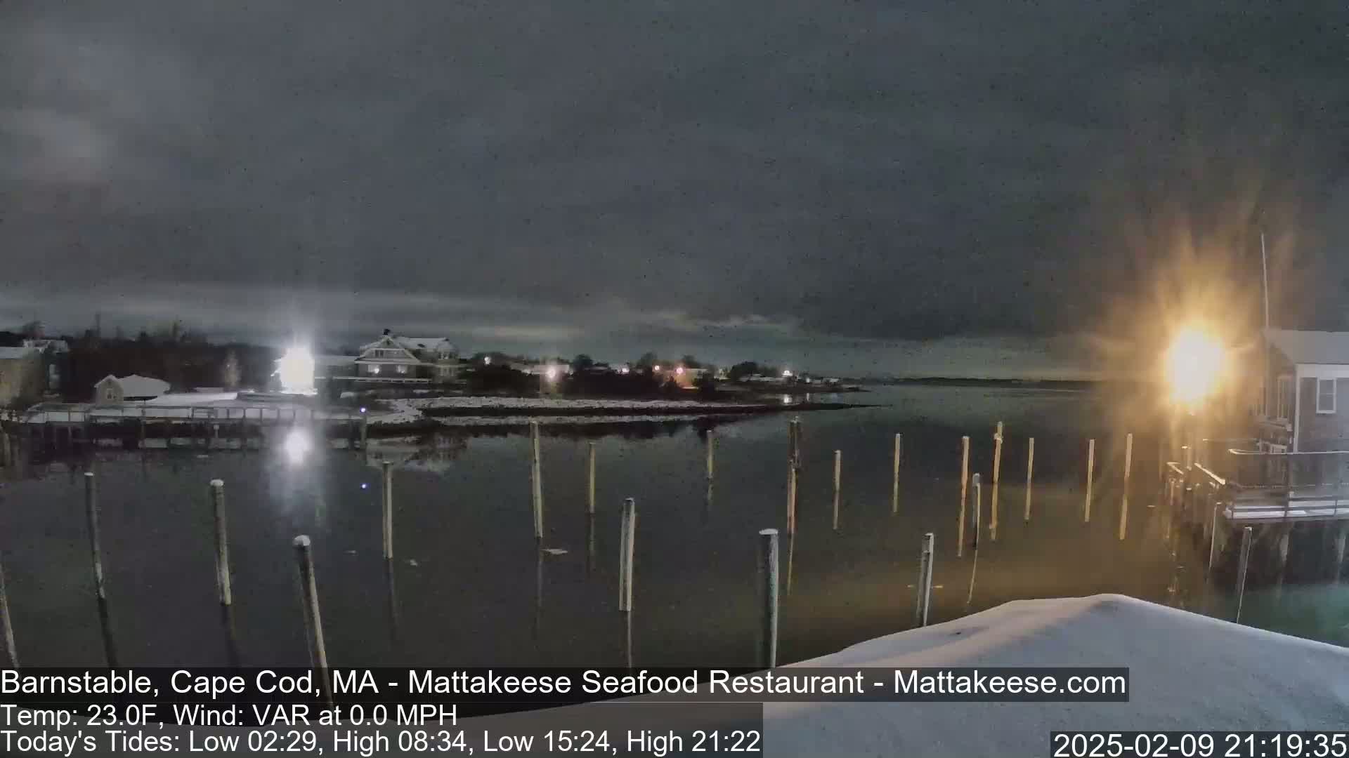 A nighttime snow scene shows a calm body of water with posts sticking up from it, a snow-covered shoreline with buildings and lights, and a dark, cloudy sky.