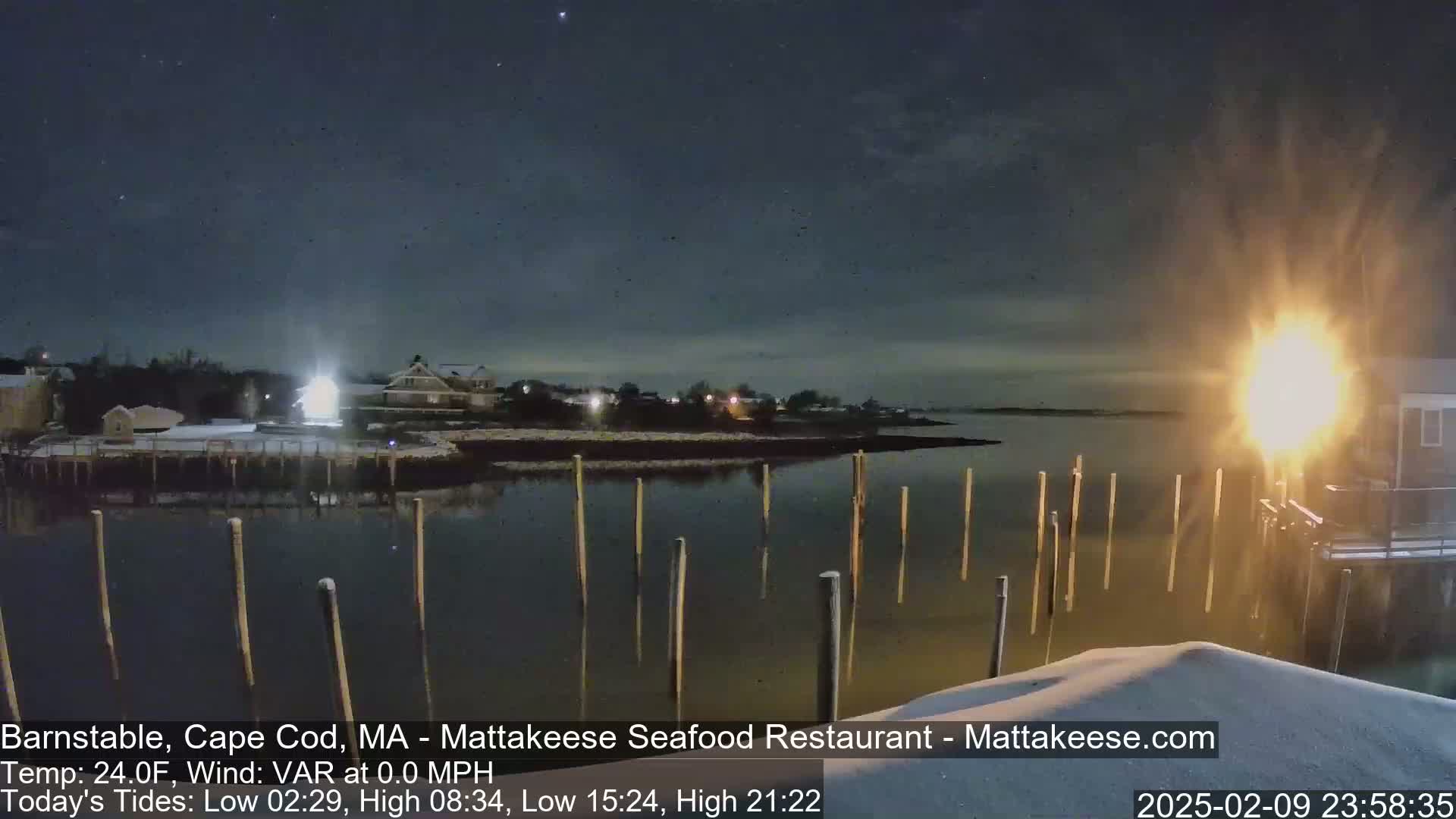 A nighttime view of a snow-covered waterfront with calm water, wooden pilings, and a few buildings lit up under a clear, dark sky.