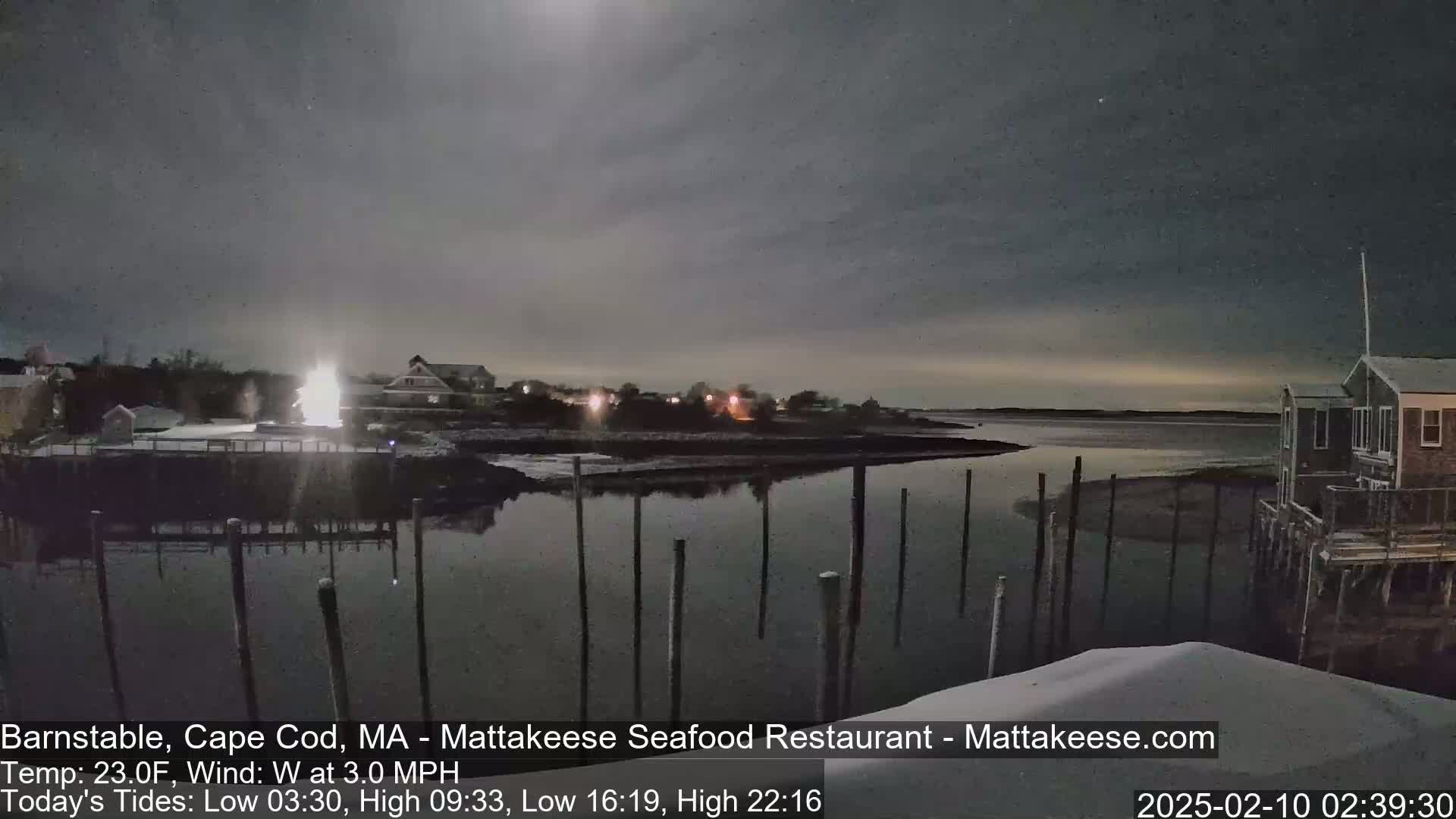 A moonlit night scene shows a calm body of water with a few houses and docks visible under a mostly cloudy sky.