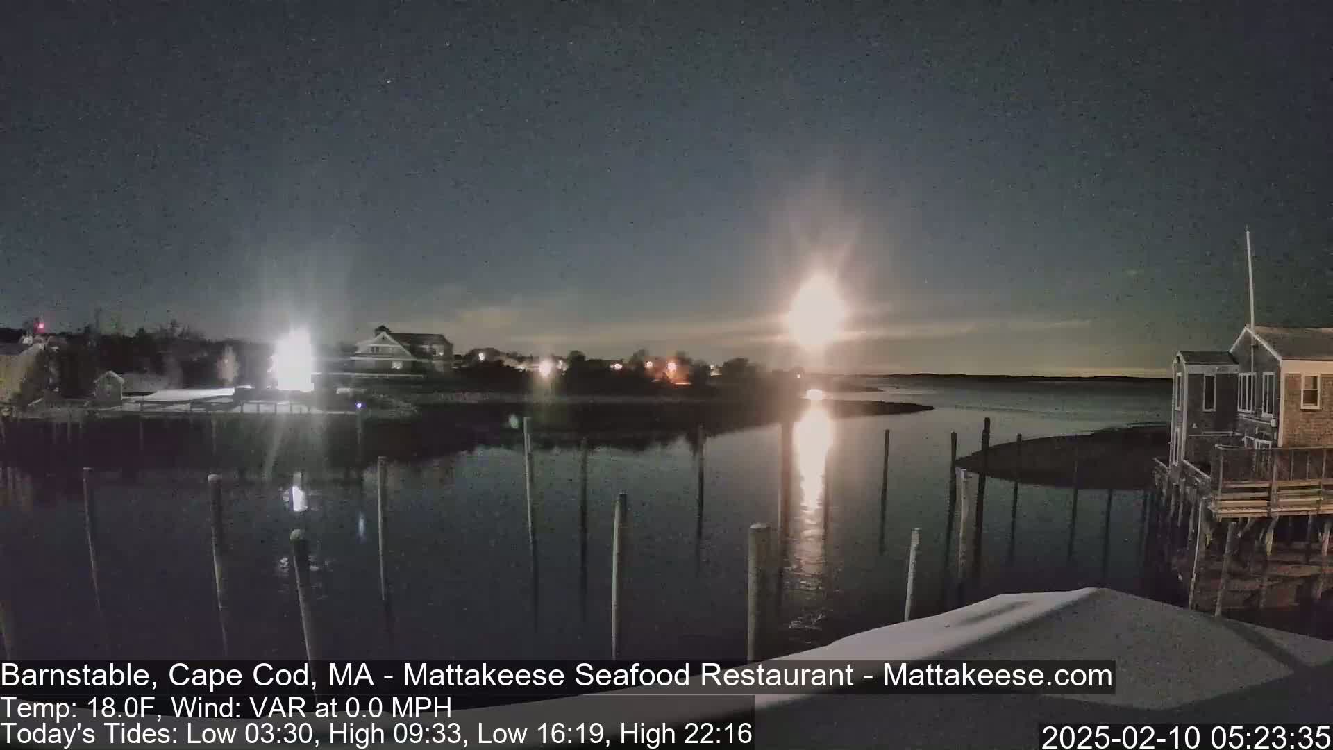 A nighttime view of a calm body of water with docks, houses, and lights along the shoreline under a mostly clear sky.
