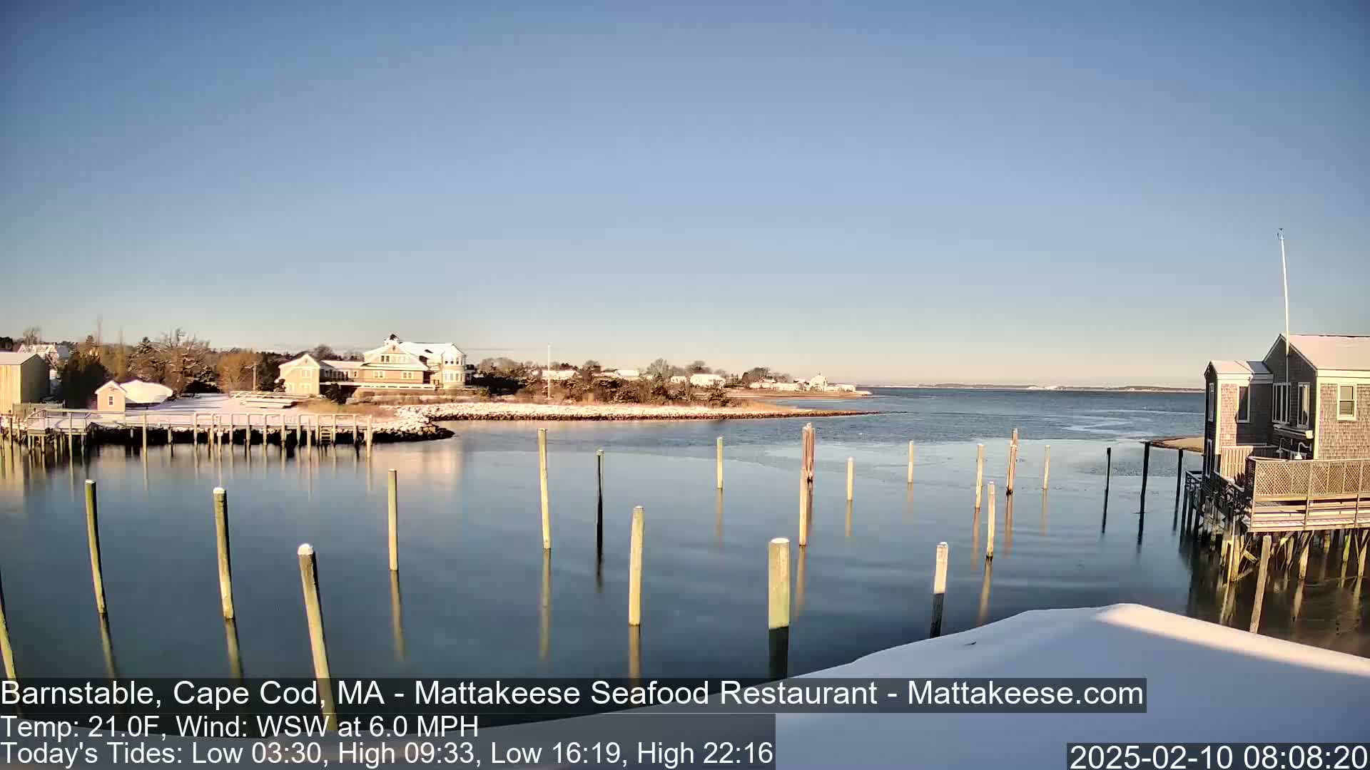 A snow-covered waterfront scene with calm water, wooden pilings, and houses under a clear blue sky.