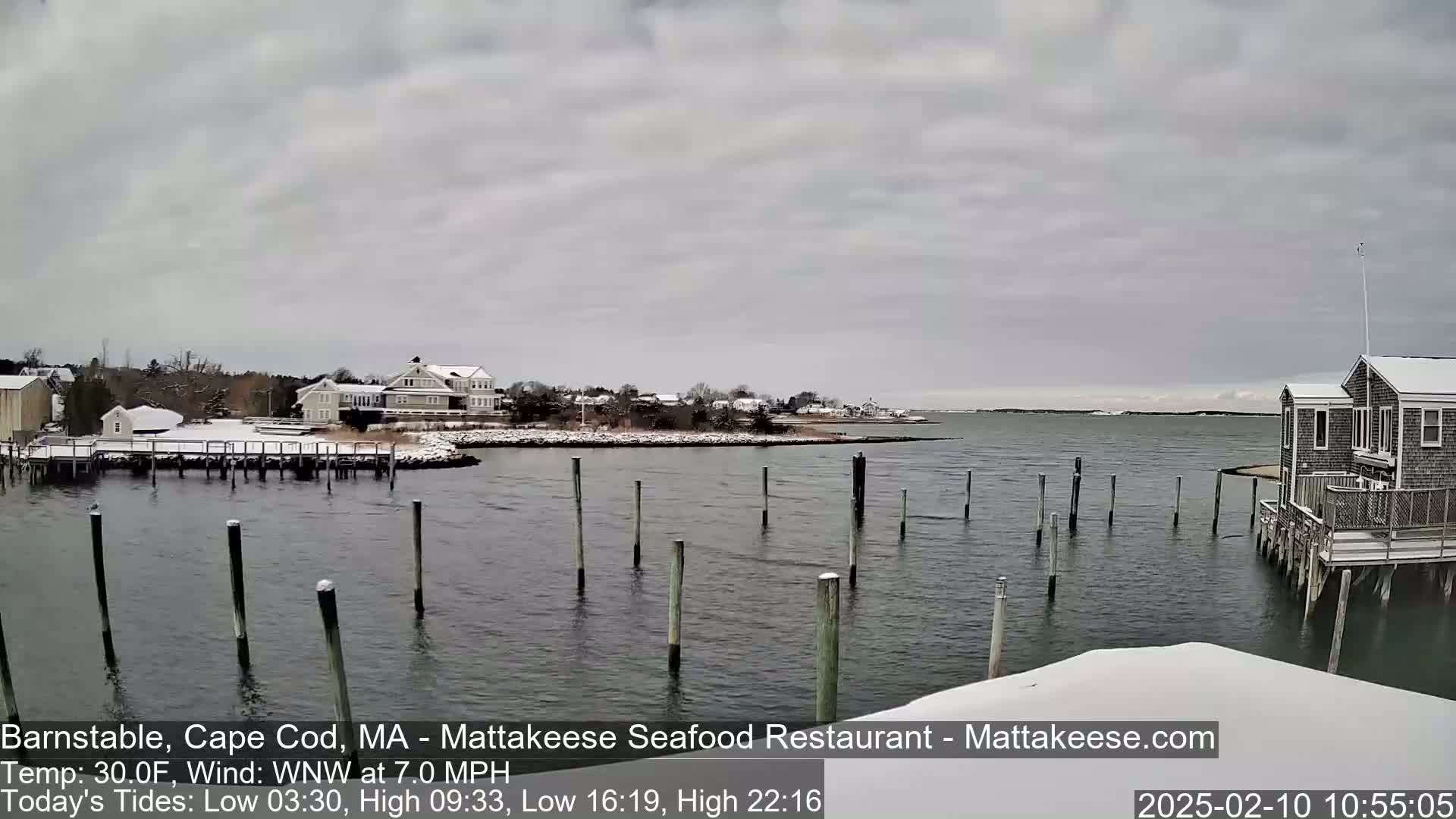 A snow-covered waterfront scene shows a calm body of water with wooden pilings, several buildings on a snow-dusted shore, and an overcast sky.