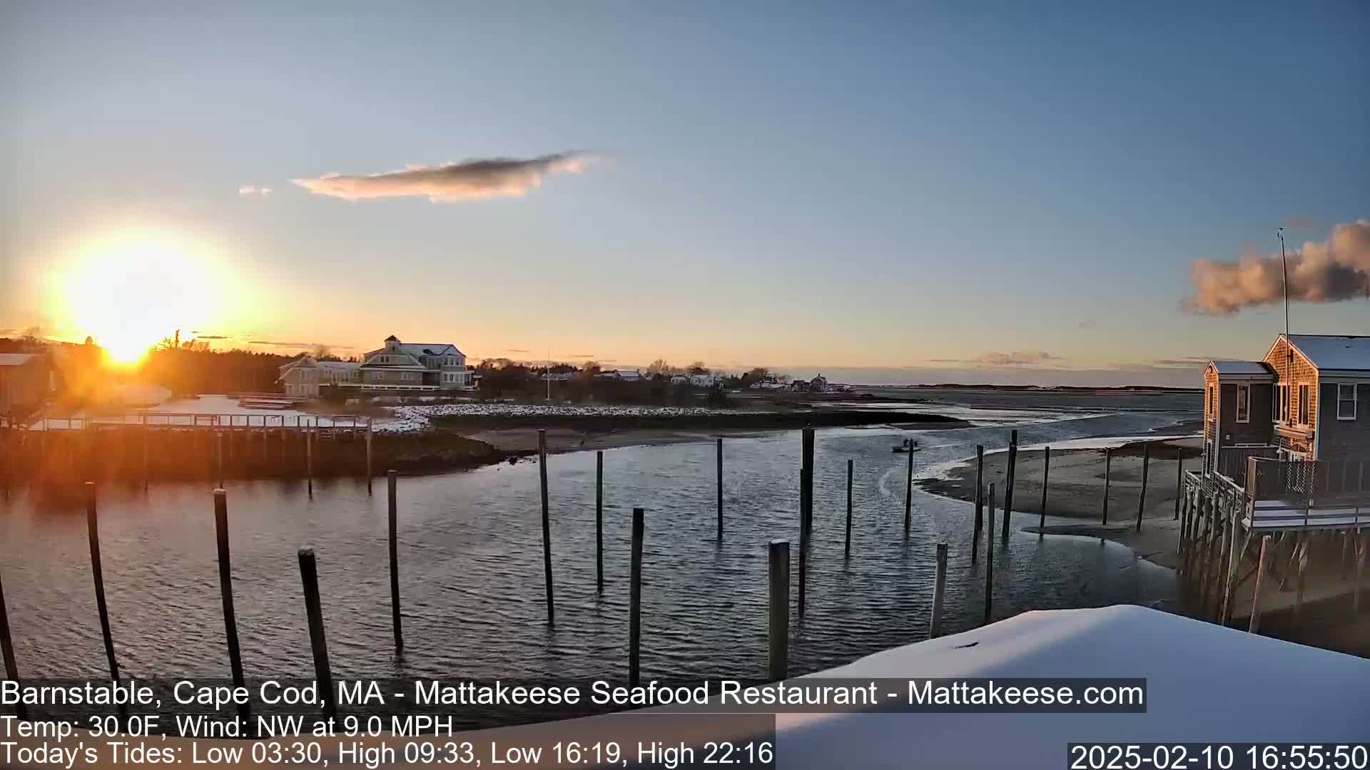 A sunset over a snow-covered waterfront scene with calm water, a few buildings, and pilings in the foreground.