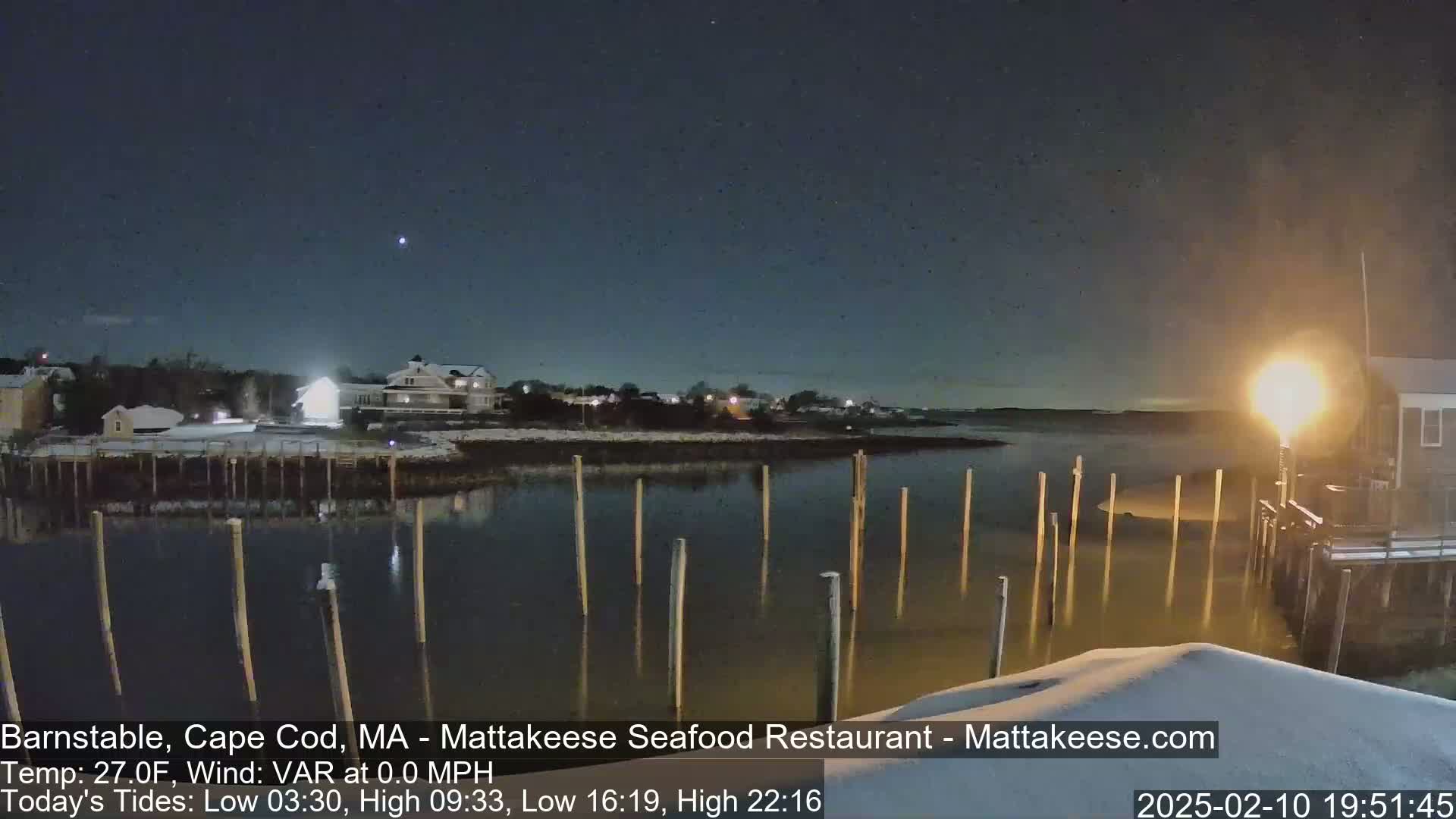 A nighttime view of a snow-covered waterfront with calm water, wooden pilings, and houses illuminated by lights under a dark sky.