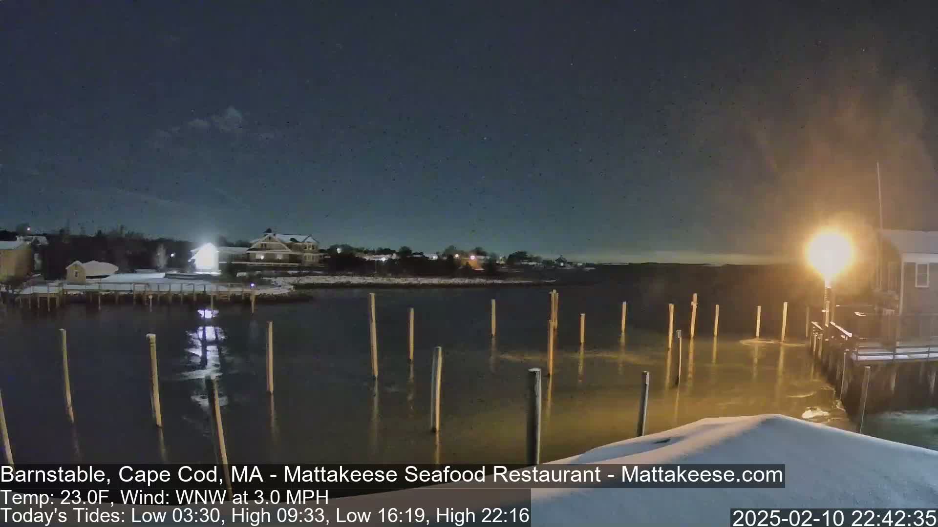 A snow-covered waterfront scene at night shows a calm body of water with wooden pilings, illuminated buildings, and a partly cloudy sky.