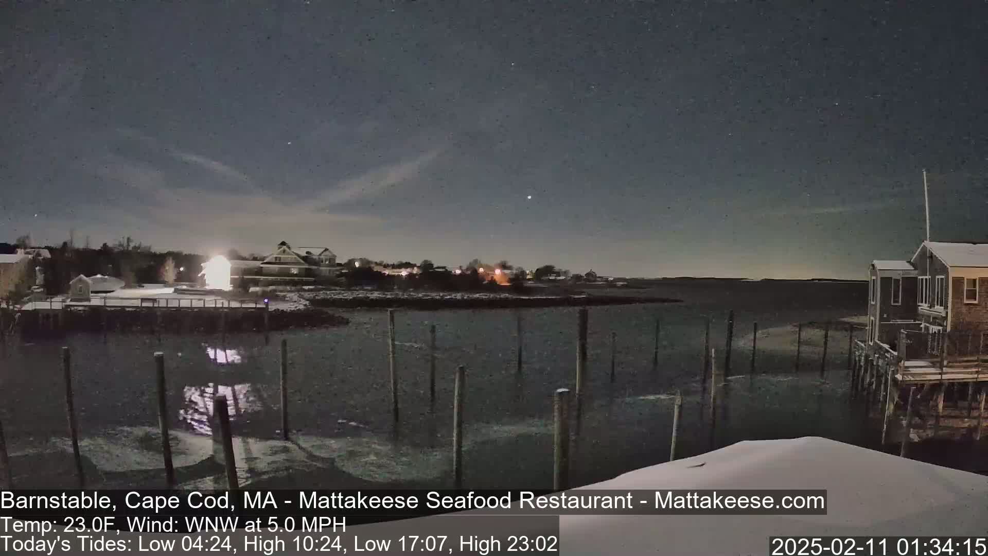 A nighttime view of a snow-covered waterfront scene with calm water, wooden pilings, and buildings under a clear, dark sky.