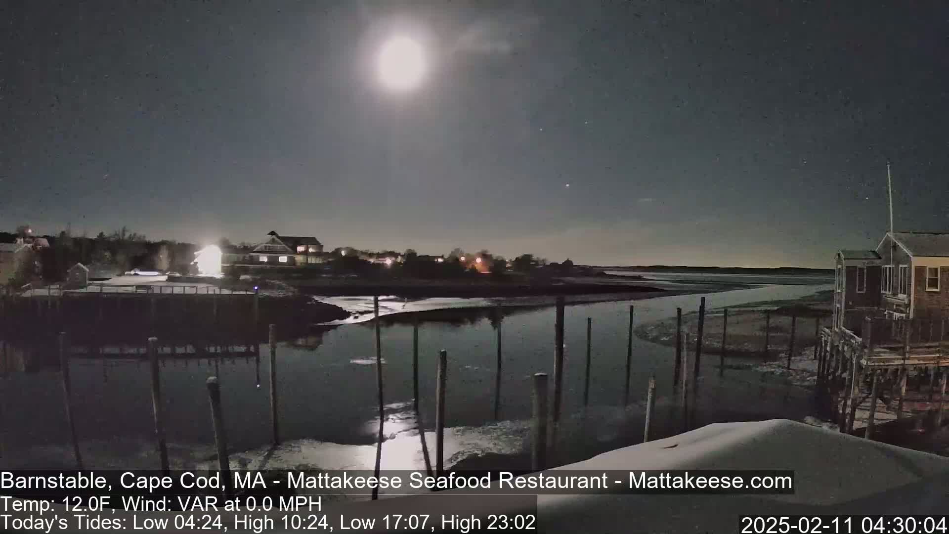 A brightly lit moon illuminates a calm waterway at night, with buildings and a shoreline visible under a clear sky.