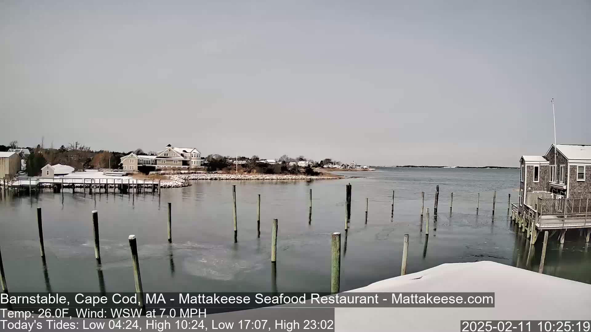 A snow-covered waterfront scene shows a calm body of water with partially frozen areas, wooden pilings, and houses on the shore under an overcast sky.