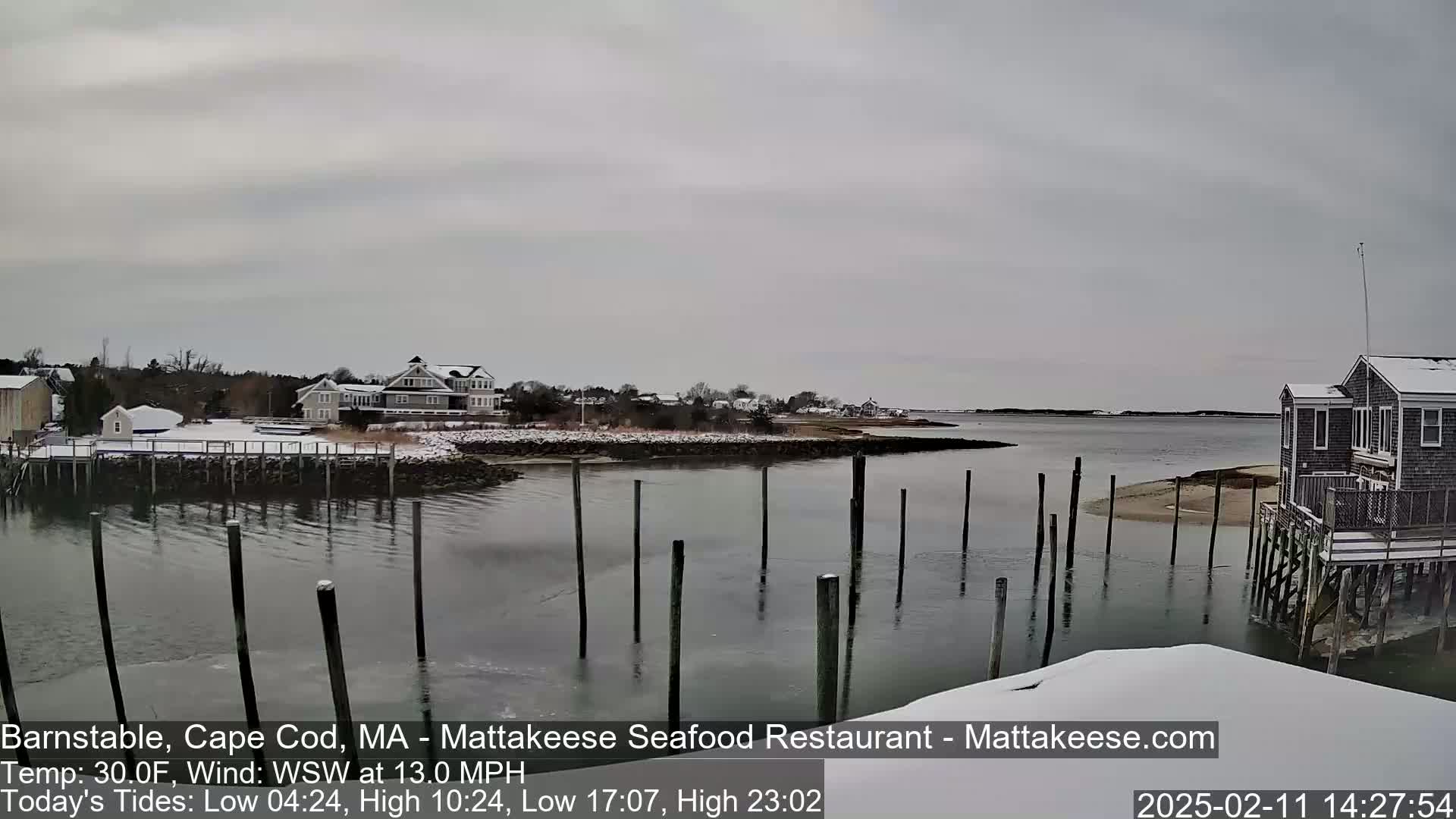 A snow-covered waterfront scene shows a calm body of water with wooden pilings, houses on a snow-dusted shore, and an overcast sky.