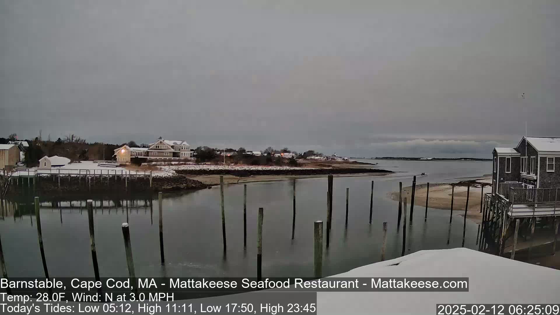 A snow-covered waterfront scene with calm water, wooden pilings, and houses under an overcast sky.
