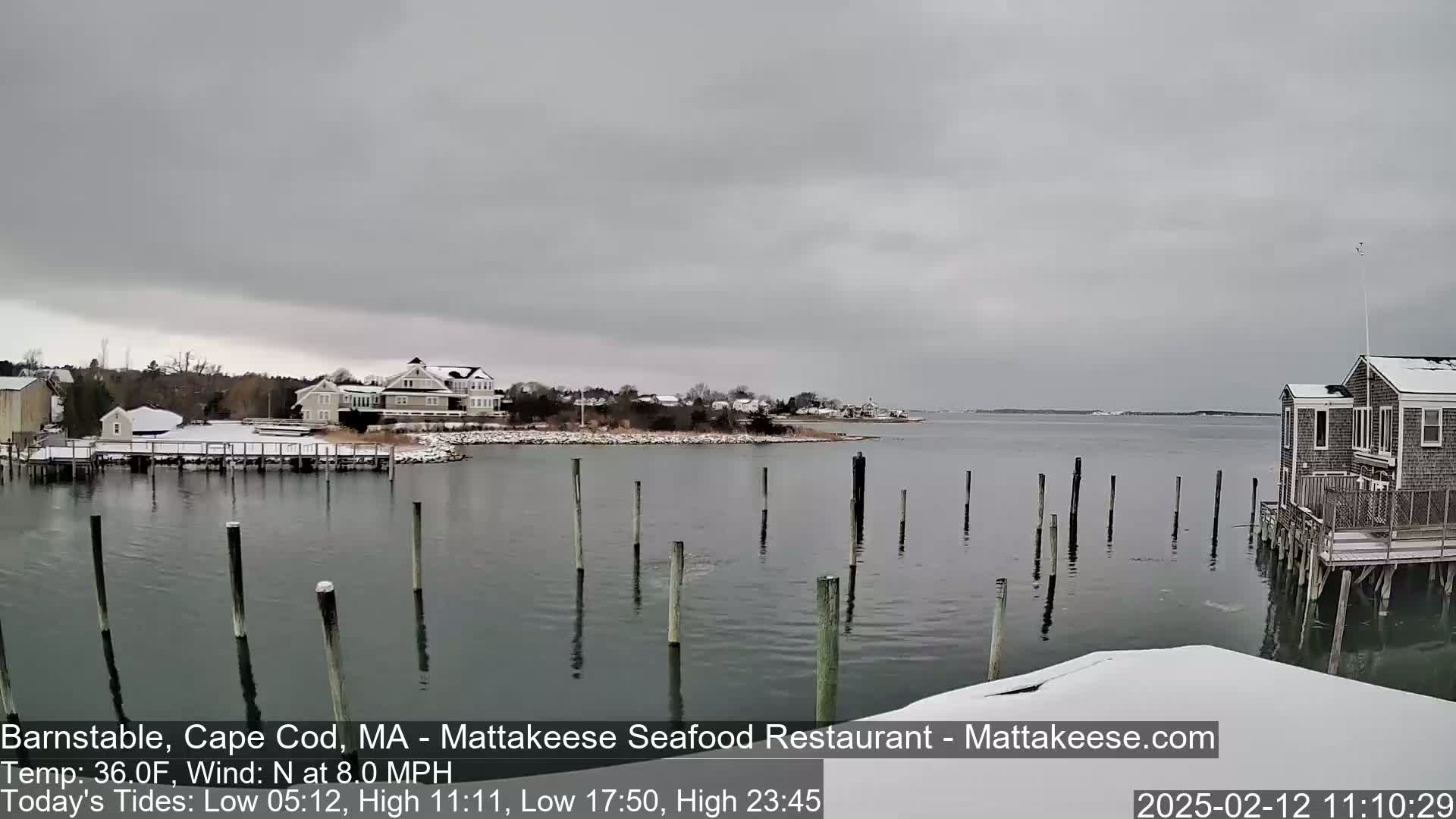 A snow-covered waterfront scene shows a calm body of water with wooden pilings, houses, and a snow-dusted shoreline under an overcast sky.
