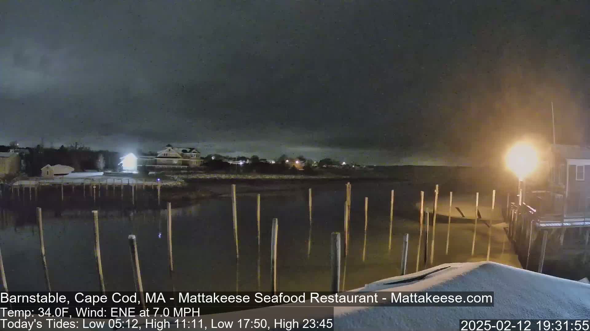 A nighttime view of a snow-covered waterfront scene with calm water and wooden pilings, under a dark, cloudy sky.